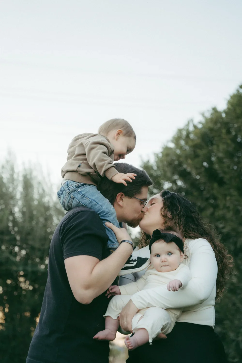 A family of four outdoors, a man and woman kissing while a young boy sits on the man's shoulders and touches the woman's head, and a baby girl is held by the woman.