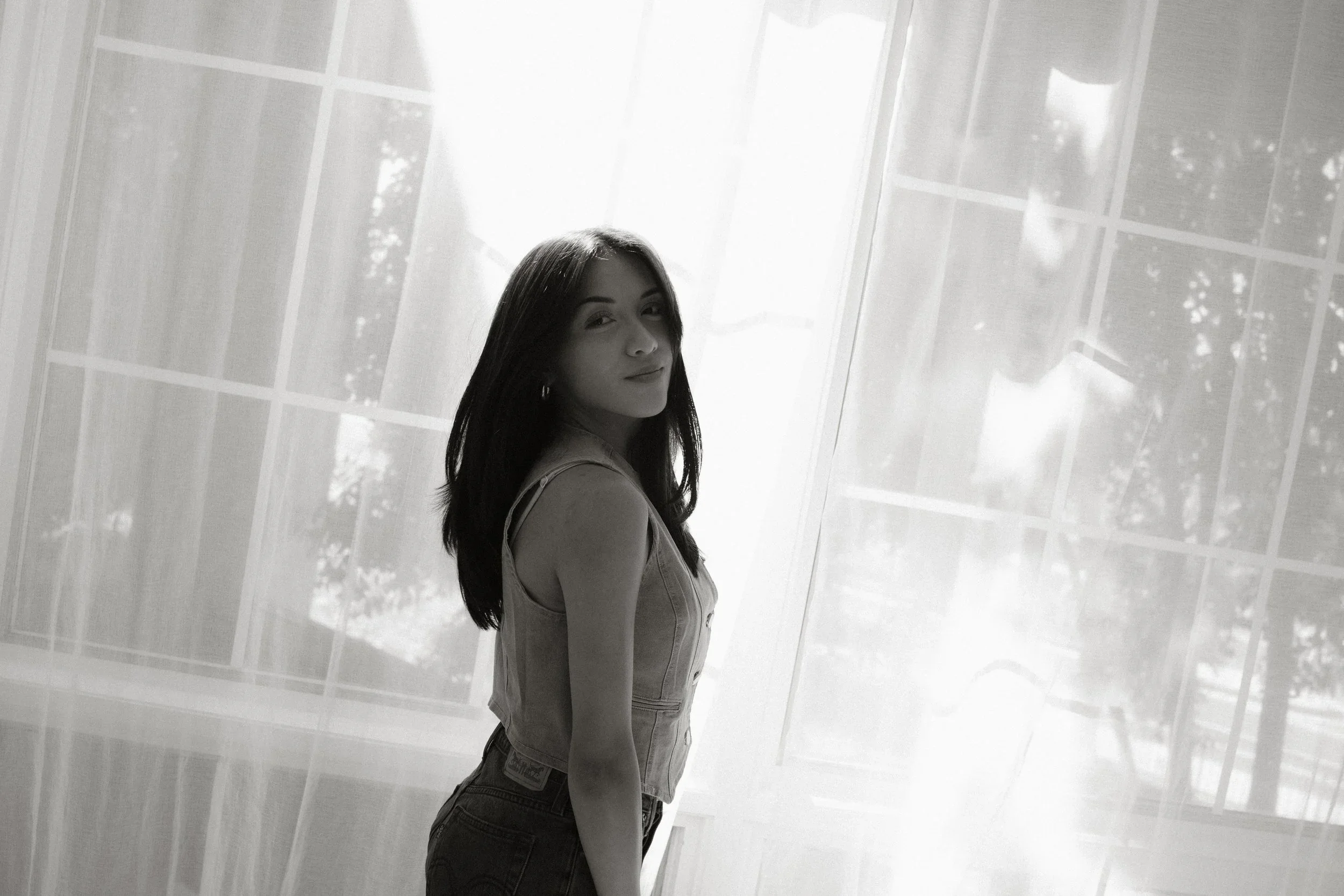 A young woman in denim overalls standing in front of a sunlit glass window, looking over her shoulder with a slight smile.