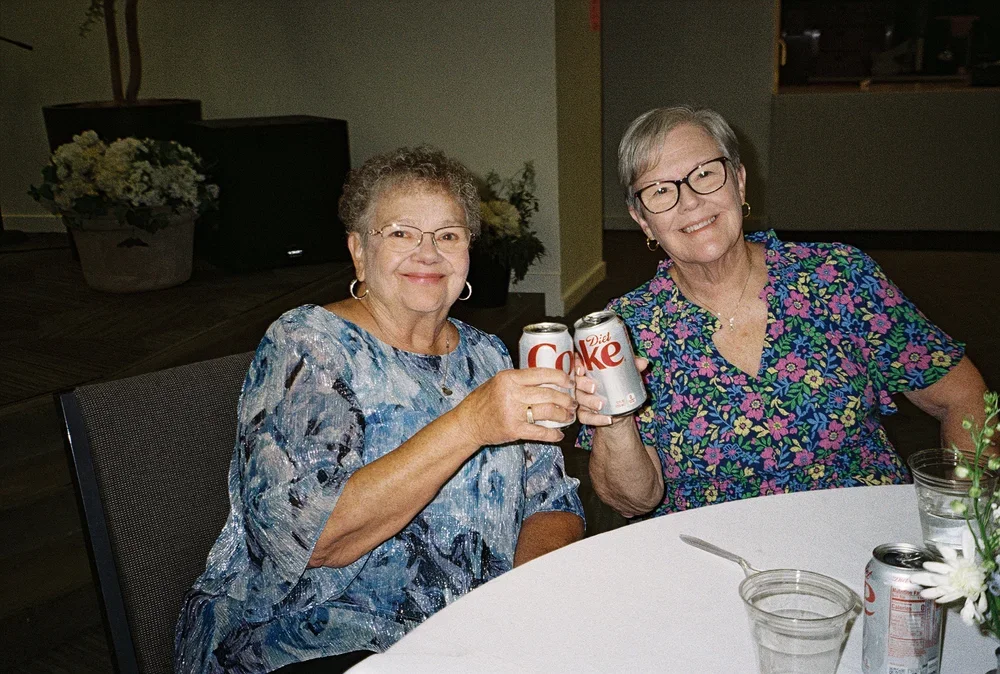 Two elderly women smiling and clinking cans of Diet Coke at a dinner table.