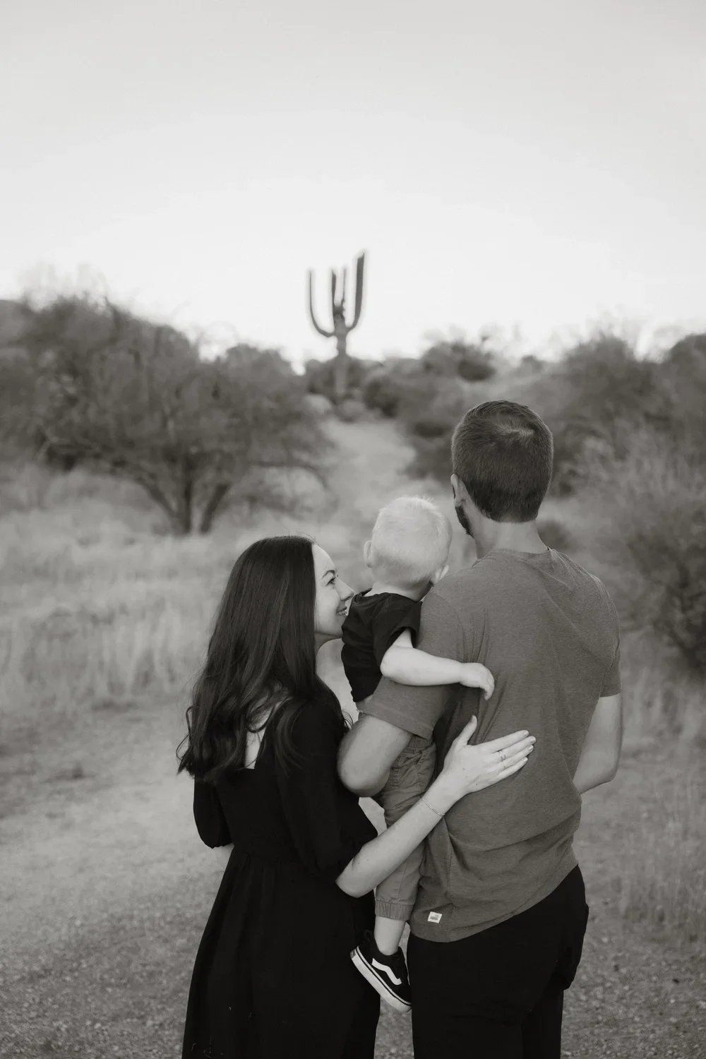 A family of three in a desert landscape with a large saguaro cactus in the background. The woman is smiling while looking at the man and holding a young boy, who is facing him. The man holds the boy with his back to the camera.