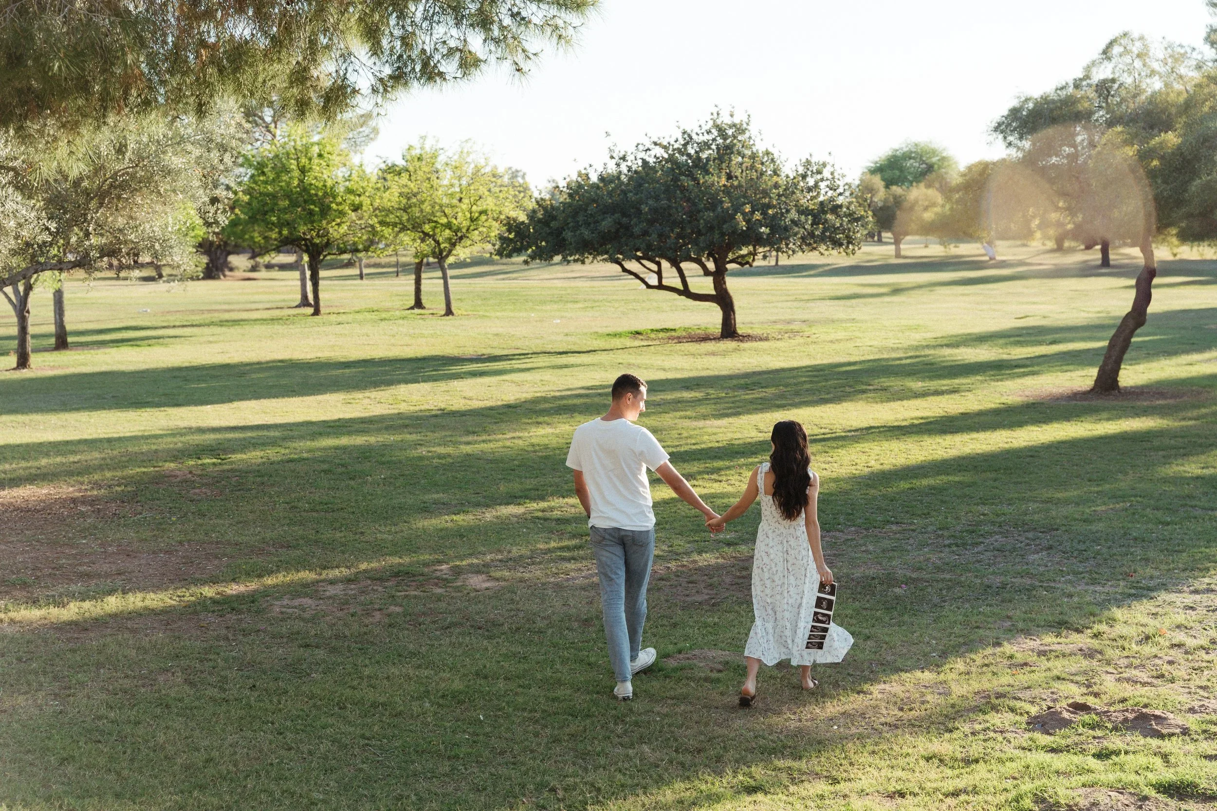 A young couple holding hands and walking in a park during daylight, with trees and grass in the background.
