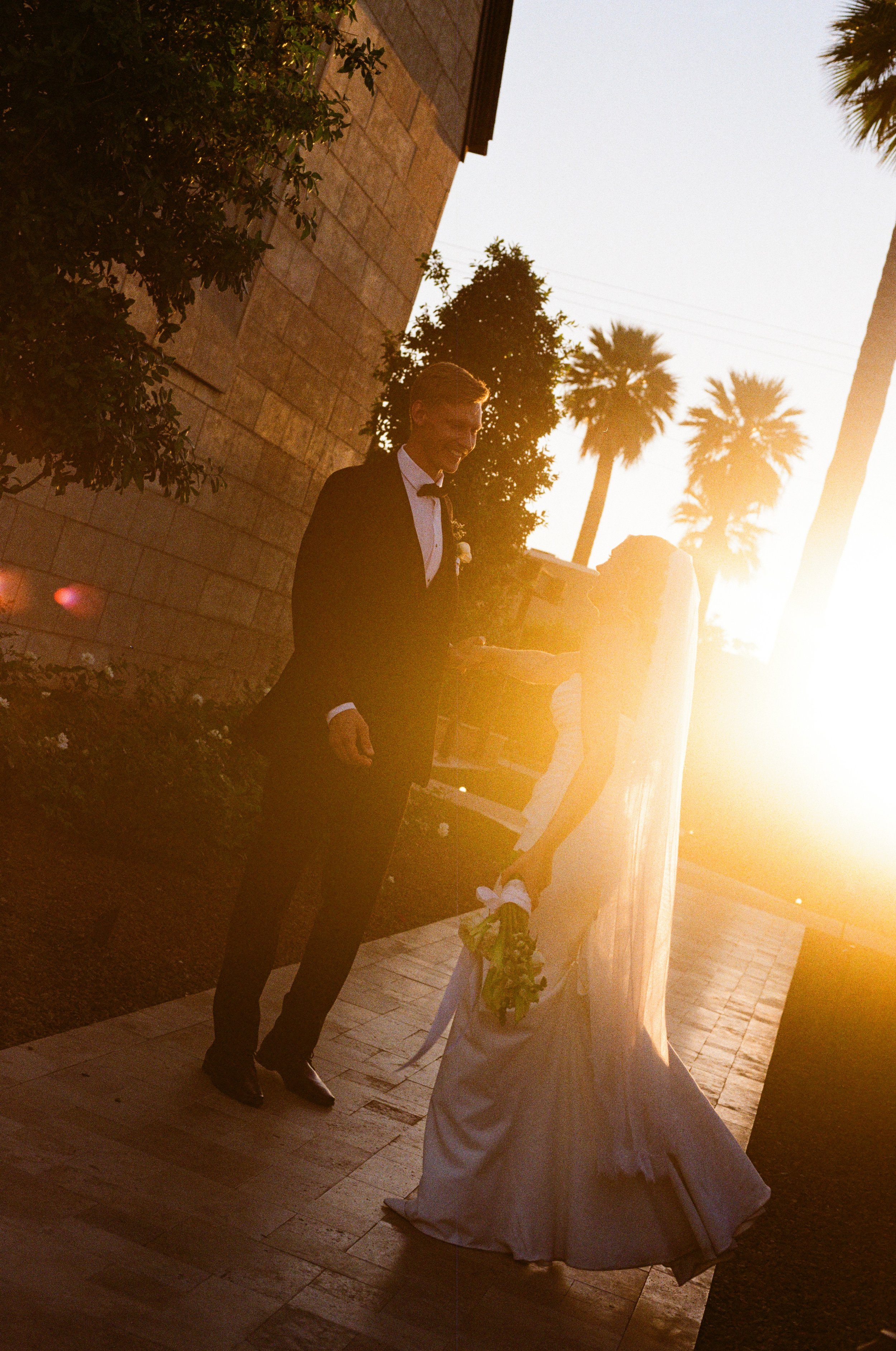 A man dressed in a tuxedo with a bow tie stands outside at sunset, smiling and looking at a bride in a white wedding gown holding a bouquet of flowers. The scene is backlit by the setting sun, creating a warm glow. Palm trees are visible in the background.
