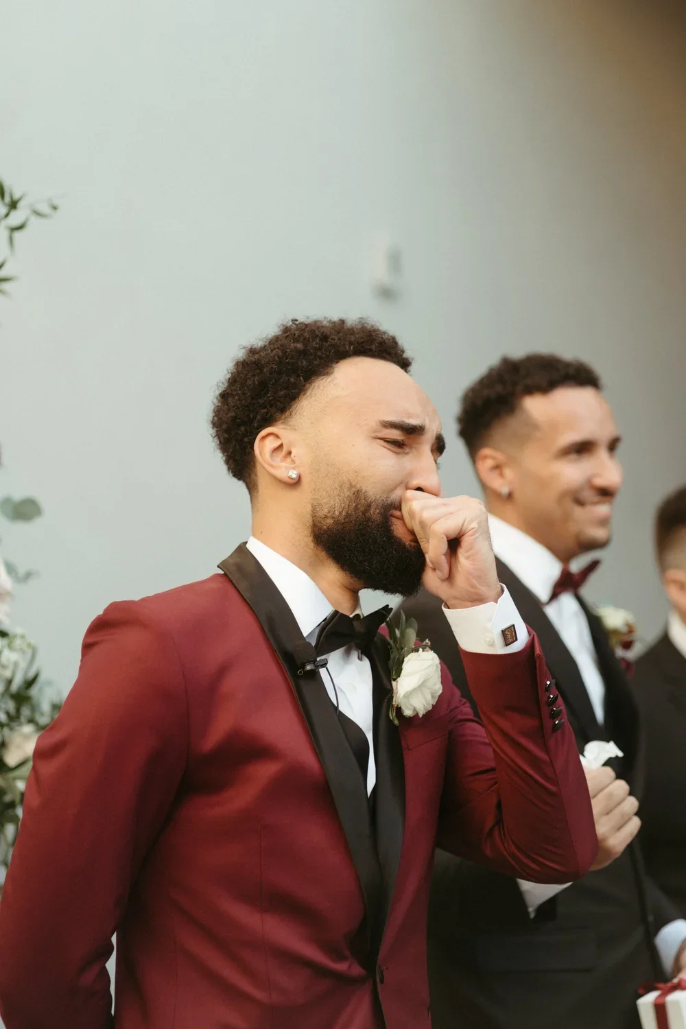 A man in a maroon tuxedo with a black satin lapel is crying during a wedding ceremony, with another man in a black tuxedo smiling beside him.