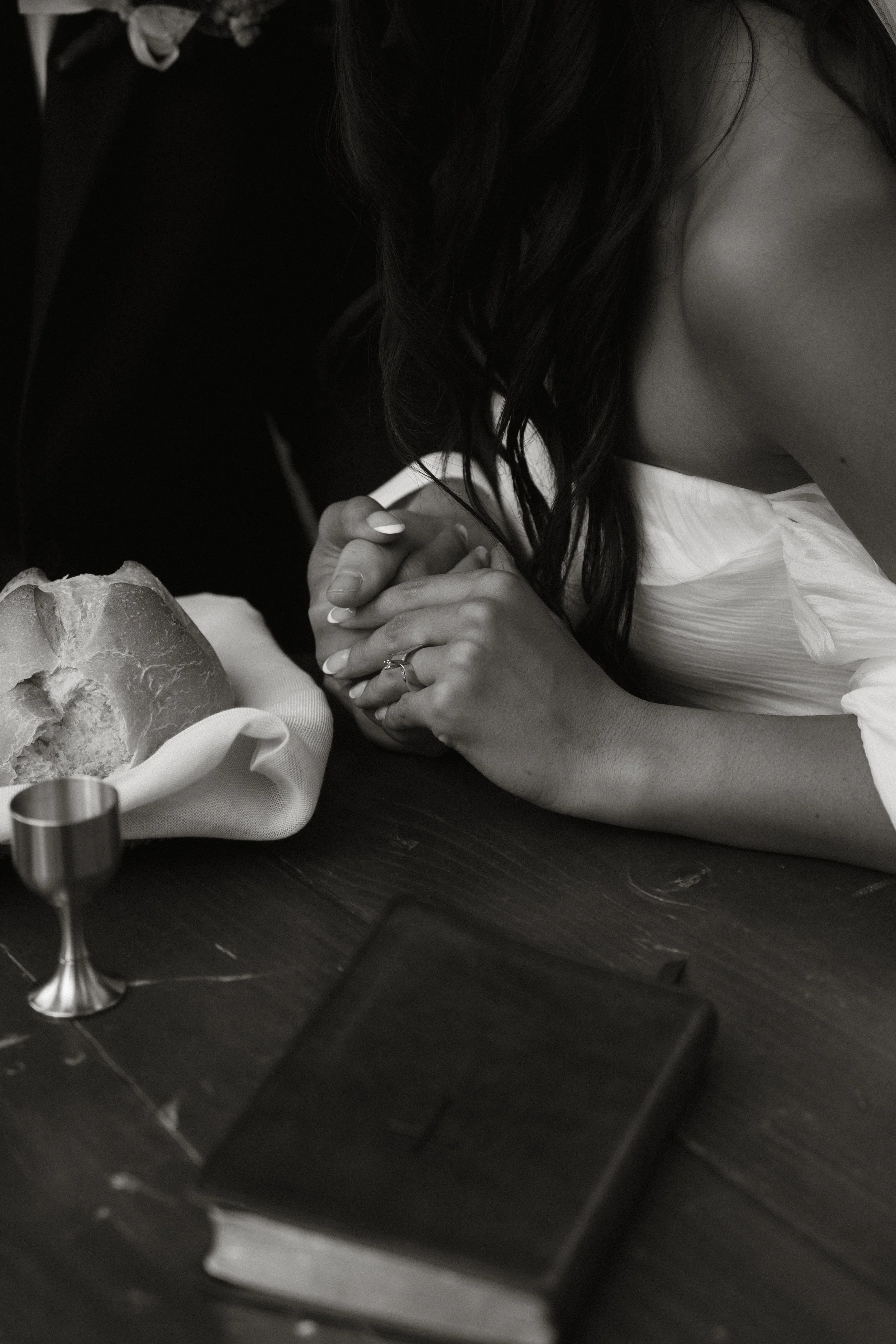 A woman with dark hair is resting her head and hands on a table, with a bread roll, a napkin, and a small silver cup nearby.