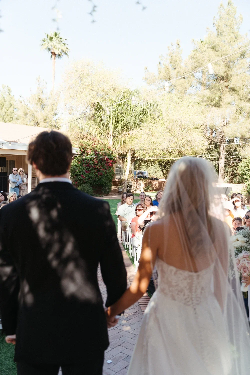 Bride and groom holding hands during an outdoor wedding ceremony, with guests seated and standing, trees in the background, under string lights.
