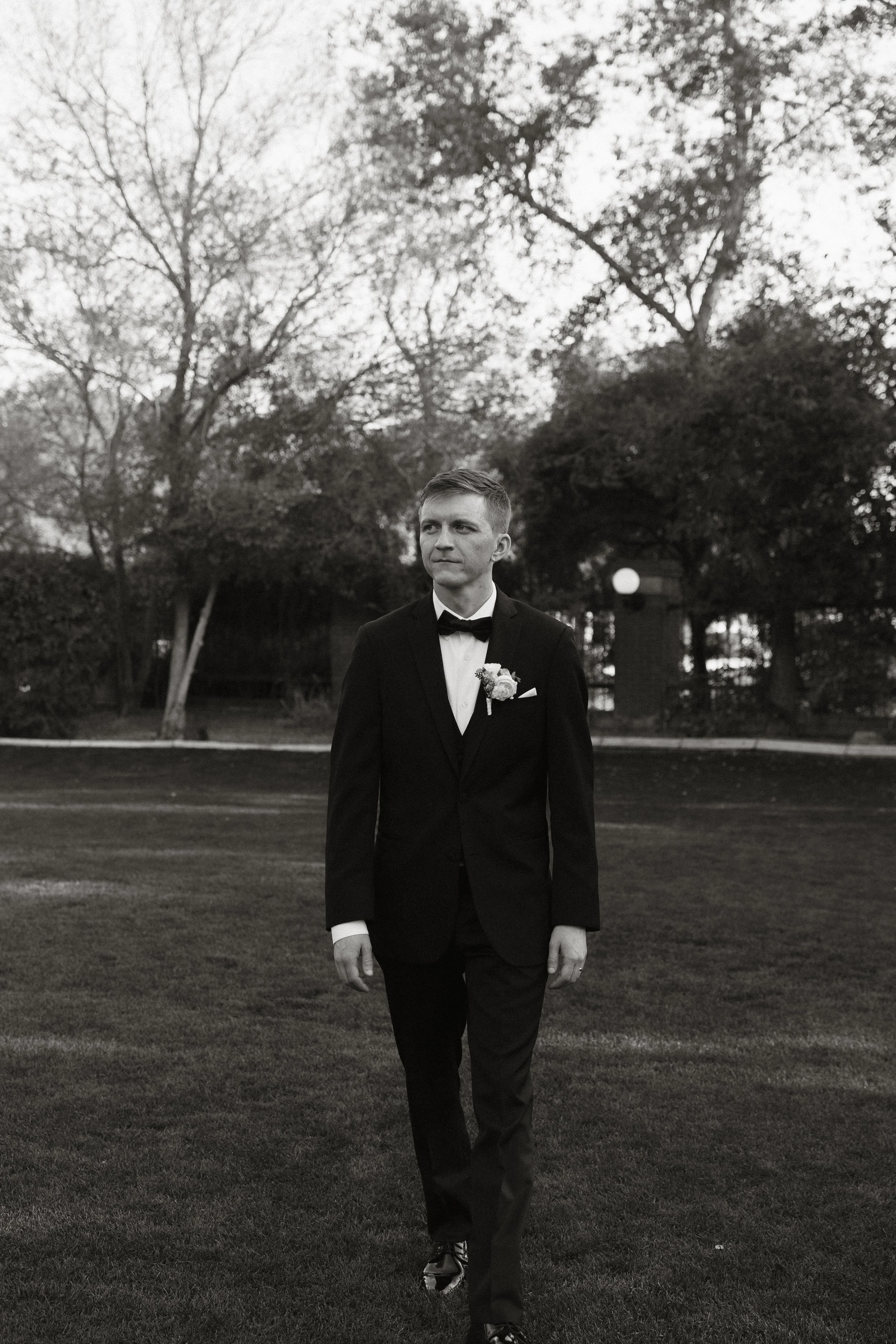 A man in a tuxedo and bow tie walking on a grassy field with trees and outdoor lighting in the background, in black and white.