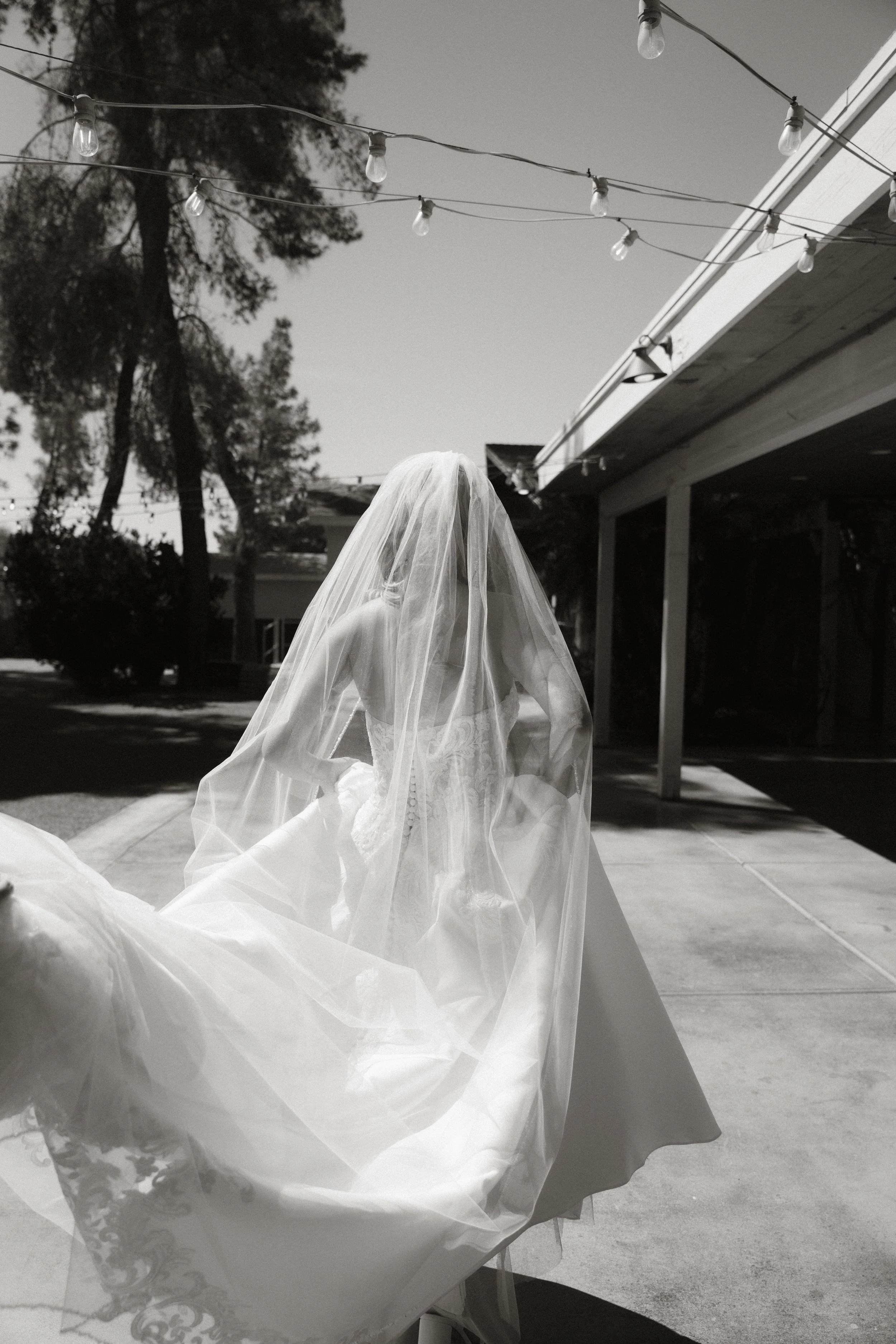A bride in a wedding dress with a veil standing outside on a paved area under string lights, with trees and a building in the background.