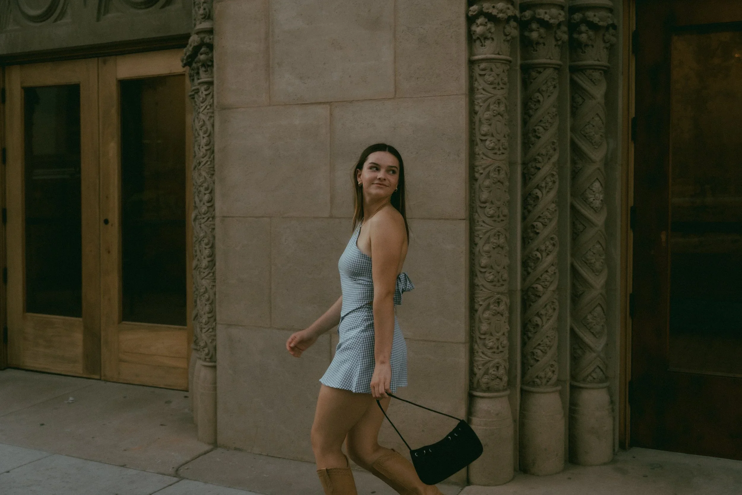 A woman in a blue and white checkered dress and brown boots walking on a city sidewalk, holding a black handbag and wearing earrings, with an ornate stone building behind her.