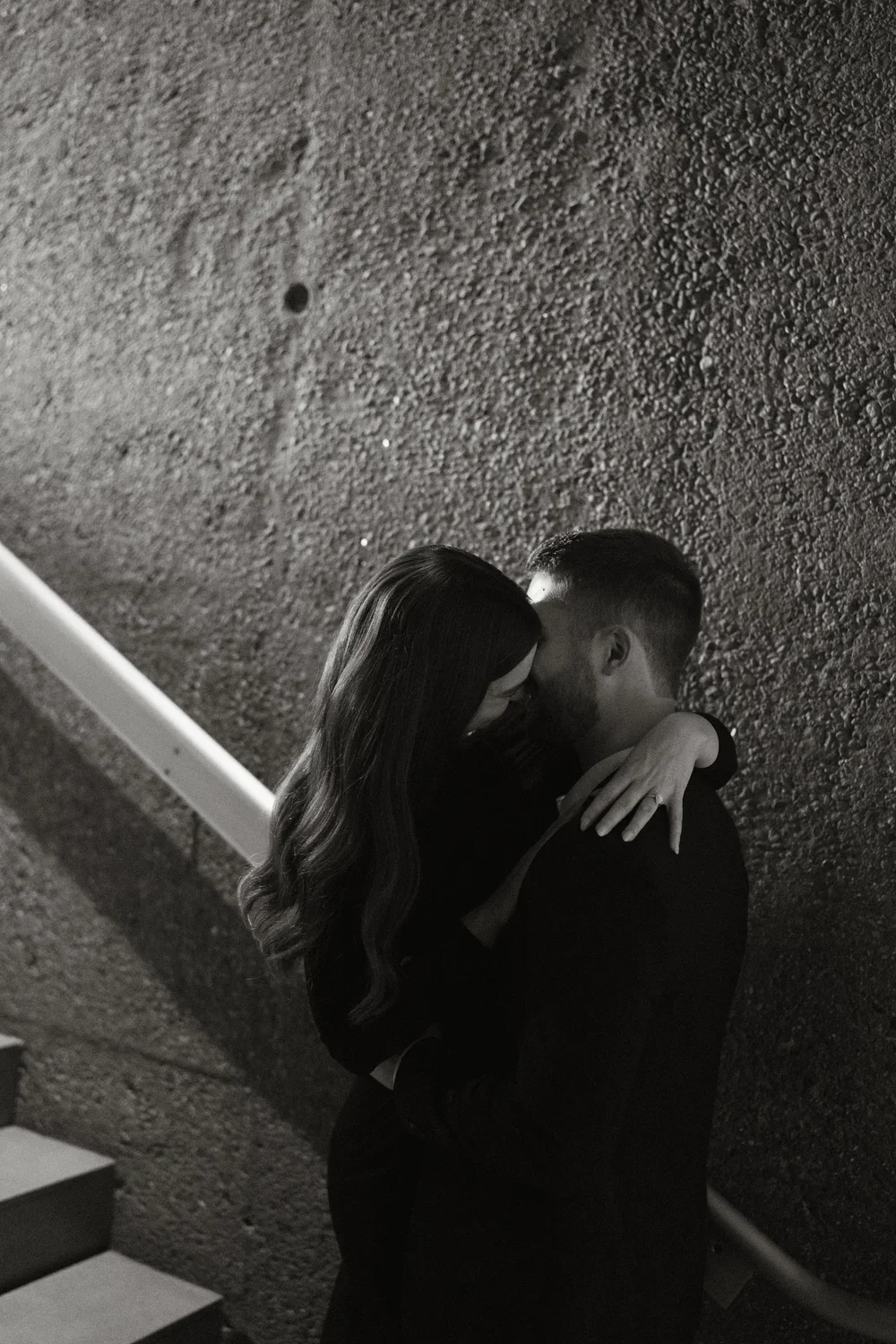 A black and white photo of a couple embracing and about to kiss, standing on stairs against a textured wall.