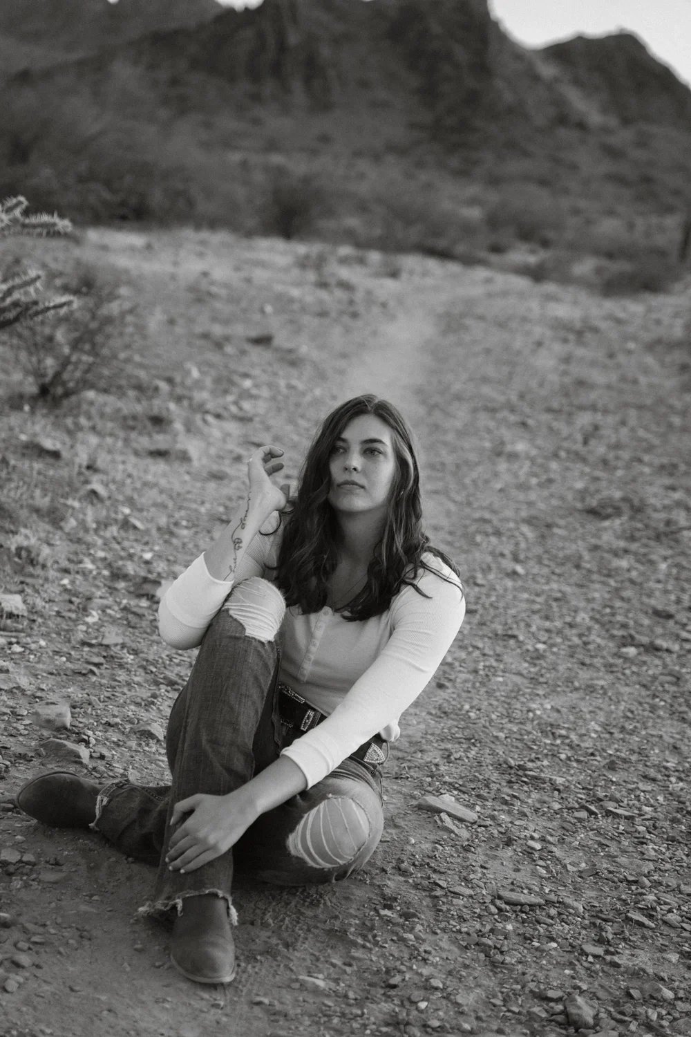 A woman sitting on desert ground with mountains in the background, in black and white.