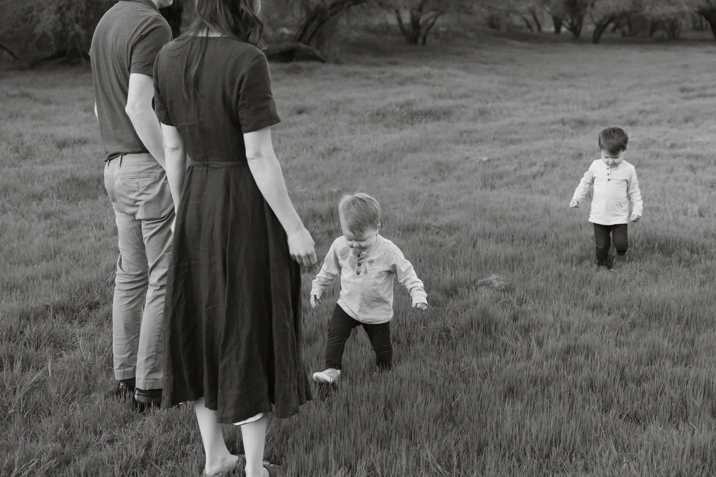 A black and white photo of a family of four outdoors in a grassy area with trees in the background. Two adults, a man and a woman, stand facing two young boys, pick up rocks or dirt from the ground.