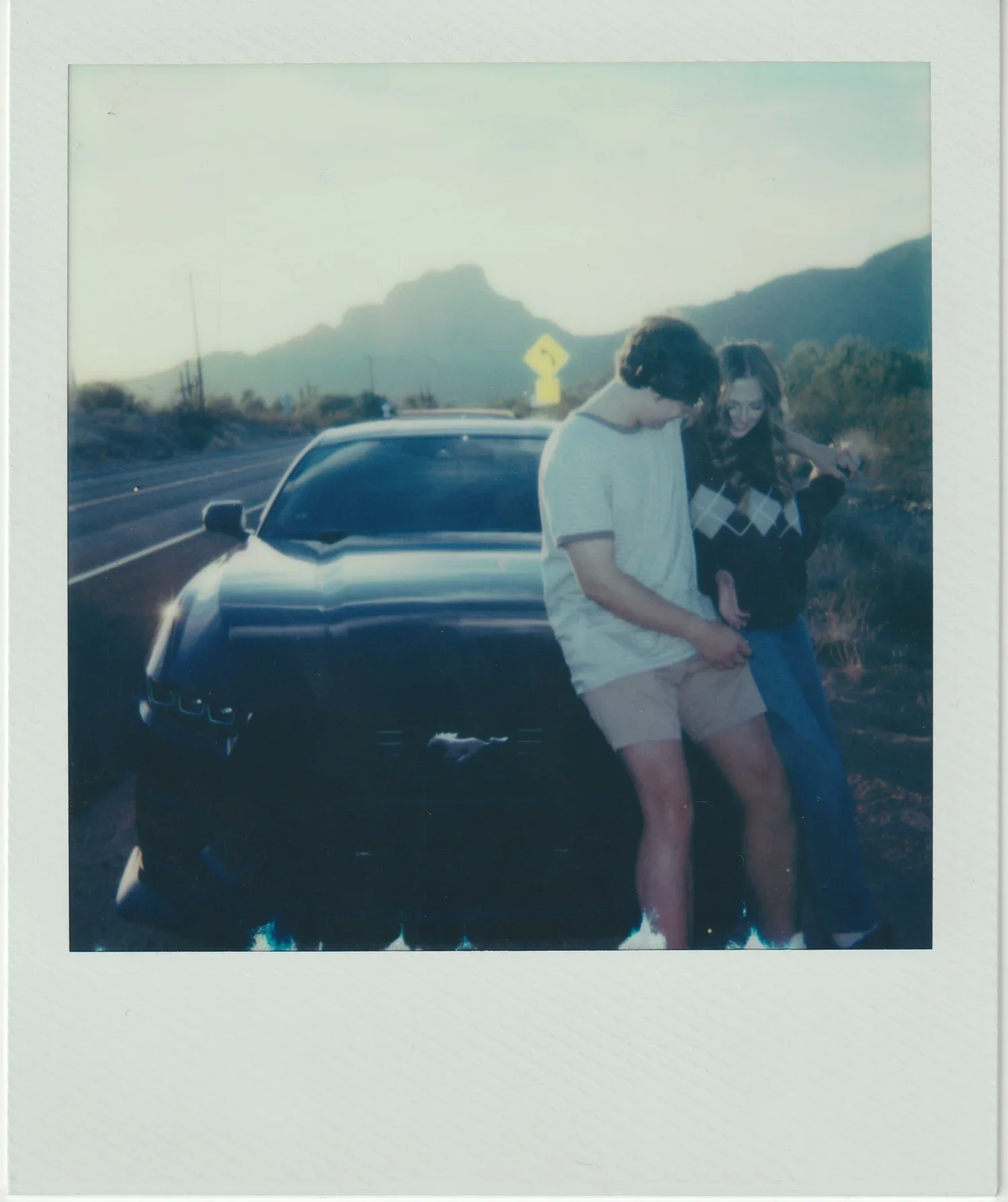 A young man and woman standing close together on the roadside, with mountains in the background and a car parked nearby during sunset or sunrise.