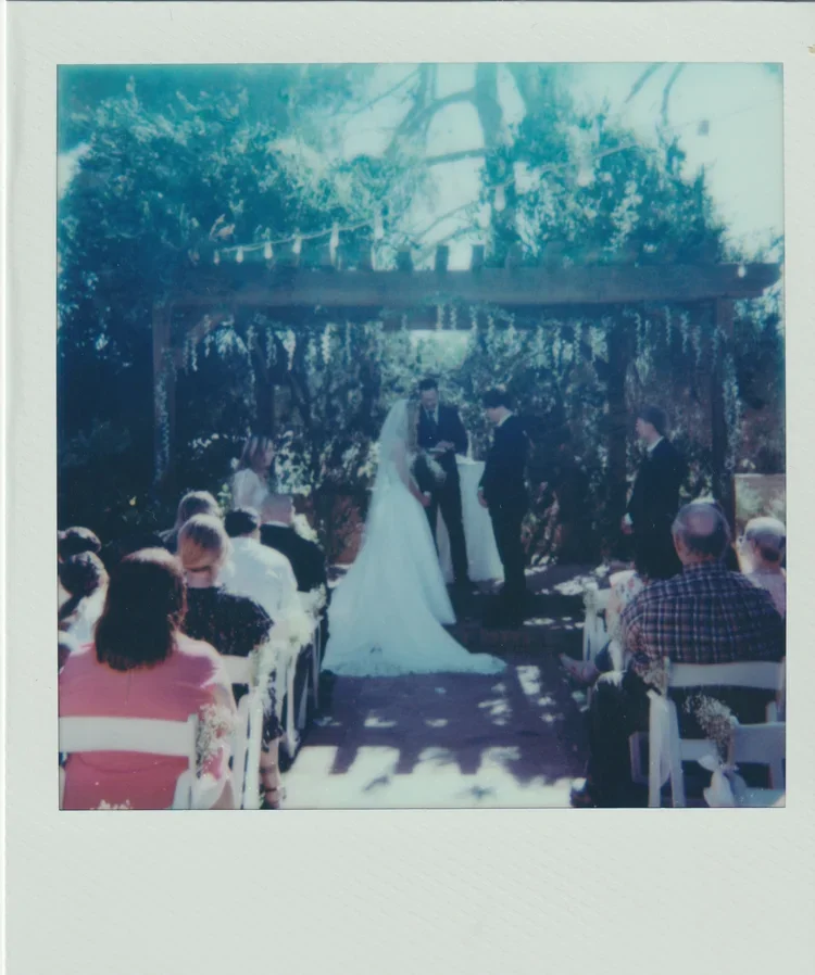 A wedding ceremony outdoors under a wooden arch decorated with greenery, with a bride and groom exchanging vows, surrounded by guests seated on white chairs.
