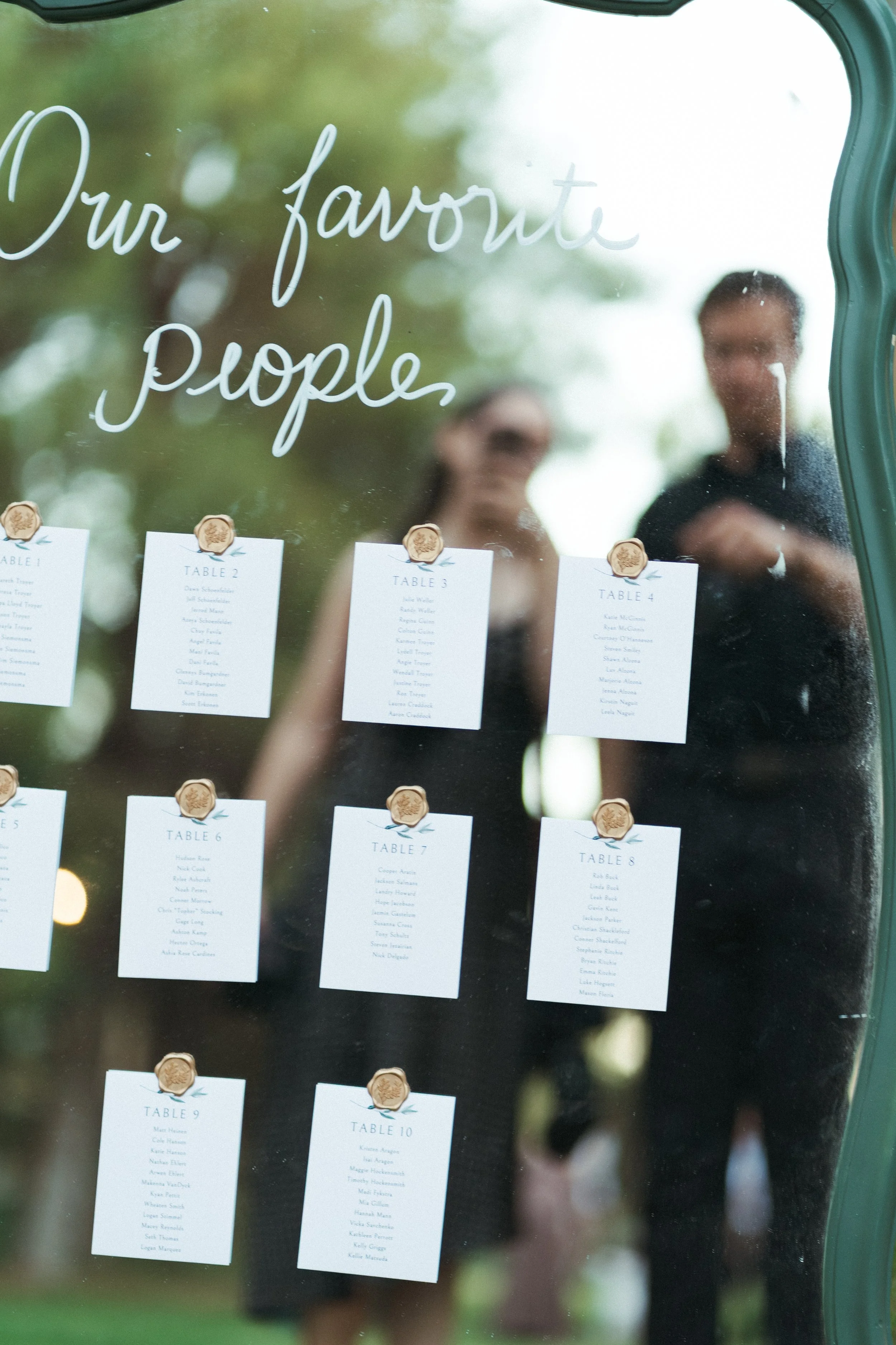 A wedding seating chart displayed on a mirror with the words 'Our favorite people' written in cursive at the top. The chart lists numbered tables with guest names under each.