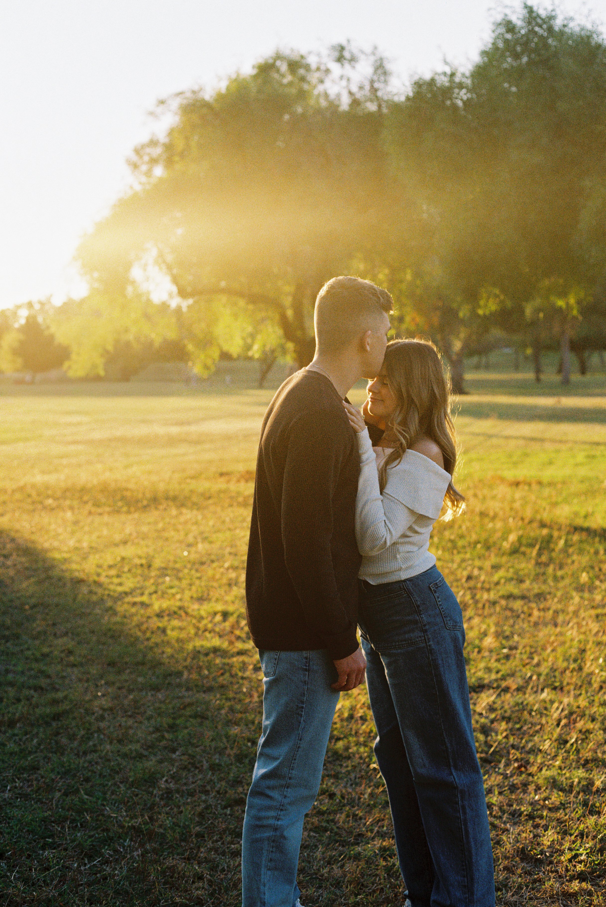 A young couple stands close together in a park during sunset, with green trees in the background. The man is facing the woman, who is smiling softly, and they appear to be sharing an intimate moment.