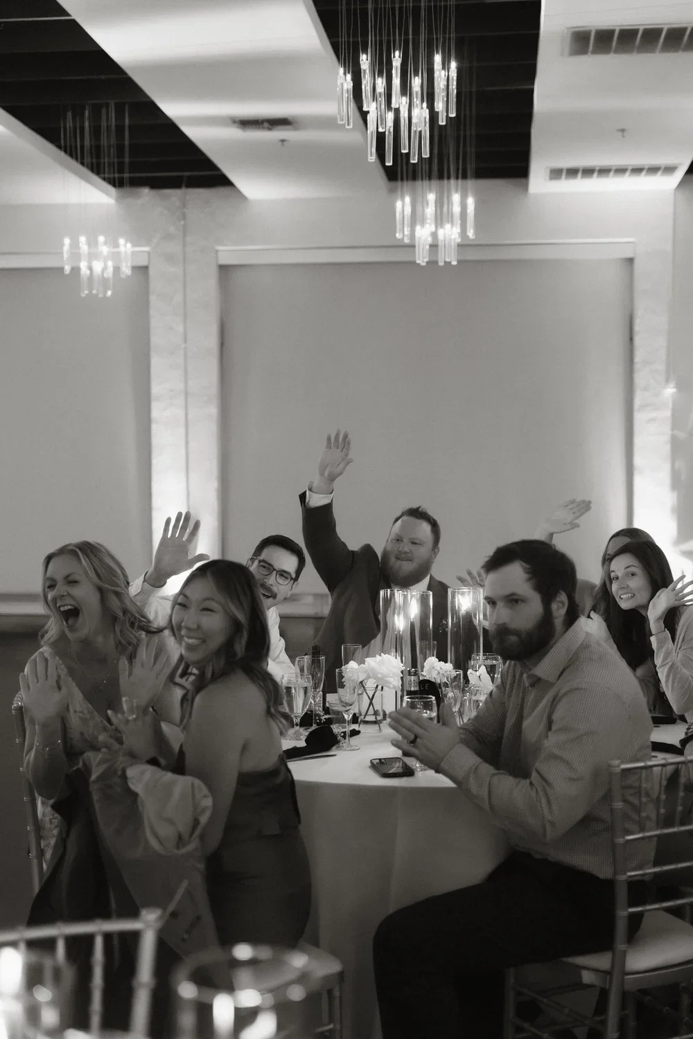 People at a formal event or celebration, sitting at tables, smiling, waving, and clapping, with elegant decor and chandeliers in the background.