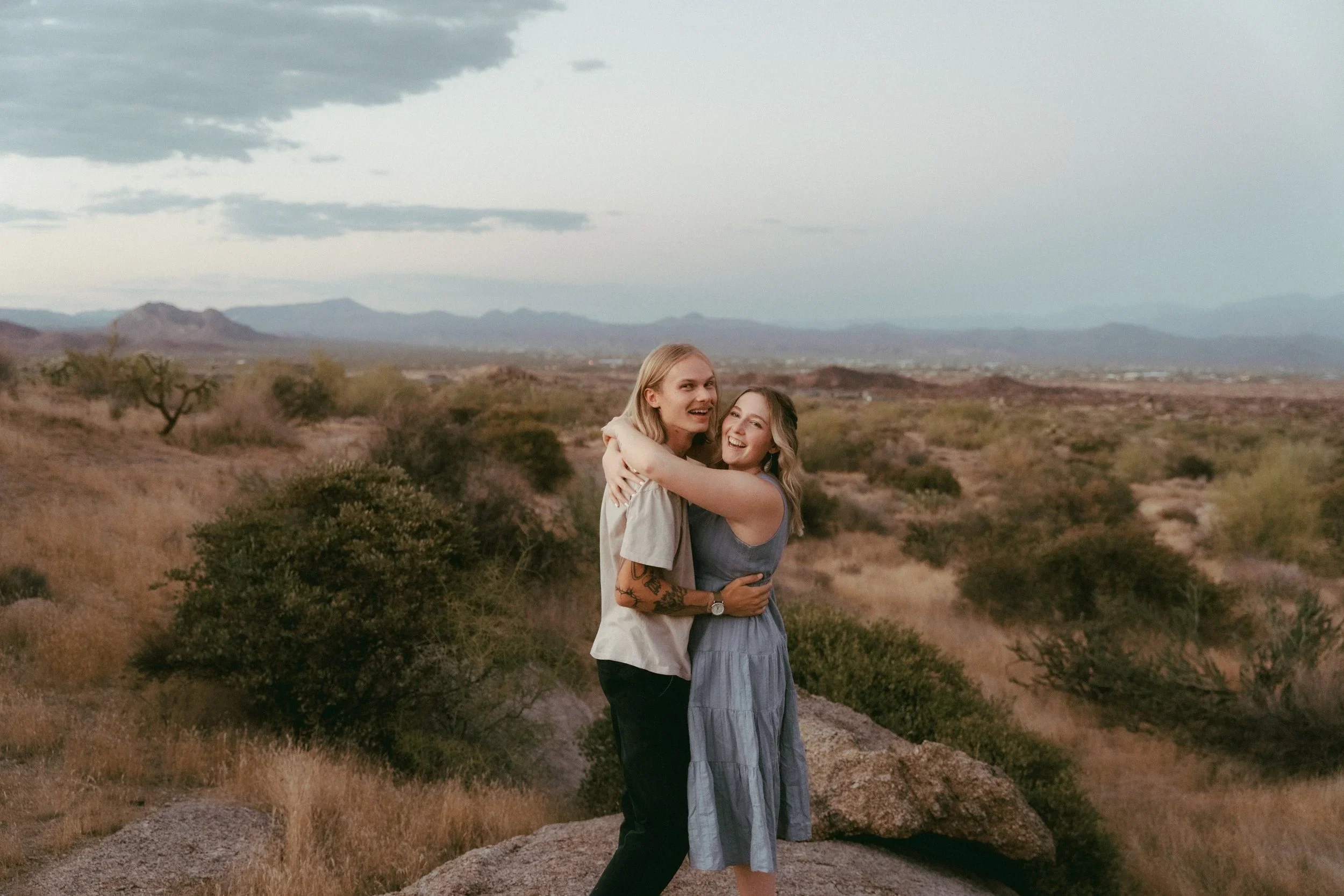 Two women embrace and smile in a desert landscape with mountains in the background, under a cloudy sky.