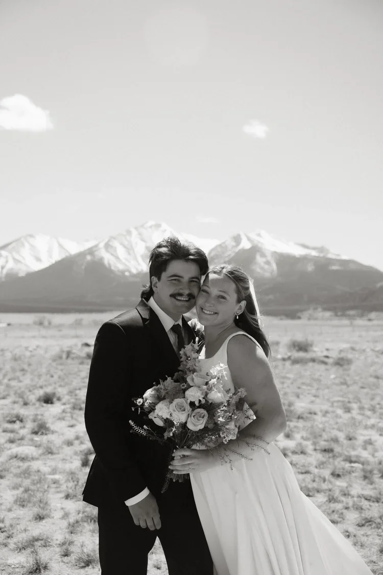 Black and white photo of a bride and groom smiling together outdoors, with snow-capped mountains in the background. The bride is holding a bouquet of flowers, and both are dressed in wedding attire.
