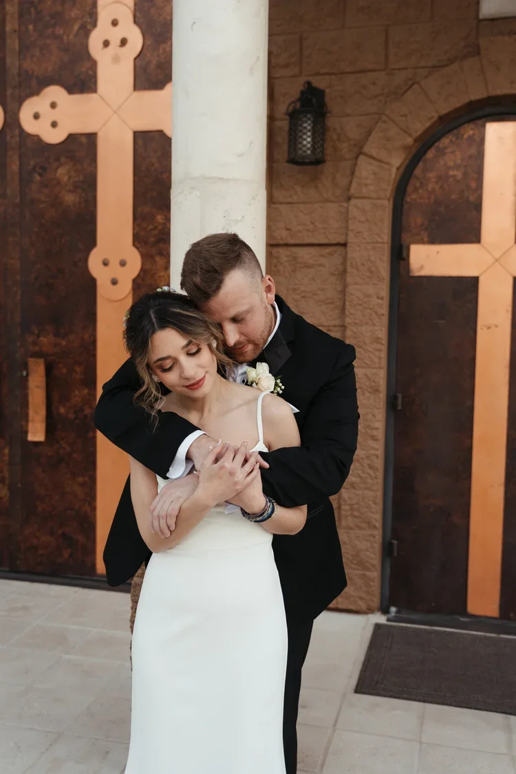 A man in a black suit hugging a woman in a white dress from behind, both with their eyes closed, in an indoor setting with brown stone walls and a decorative cross on the wall.