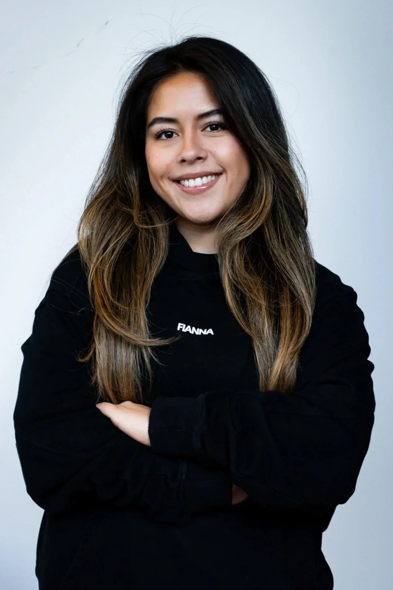 A smiling woman with long wavy brown hair, wearing a black shirt that says 'FIANNA', standing in front of a dark background with a Ford logo.