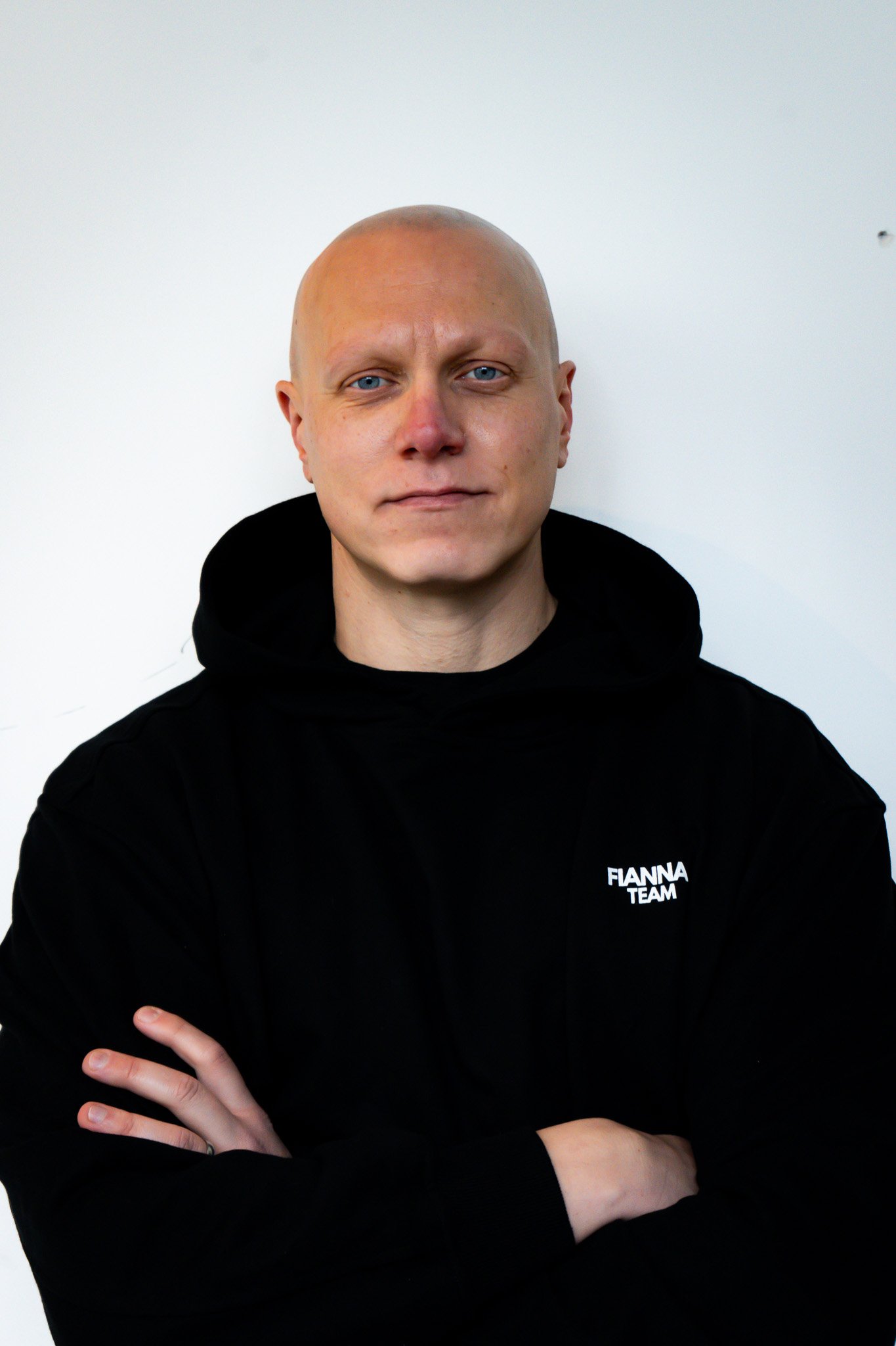 A man with light skin and a shaved head smiling with arms crossed, wearing a black T-shirt with the word 'FIANNA' on it, standing against a dark, vertically-lined background.