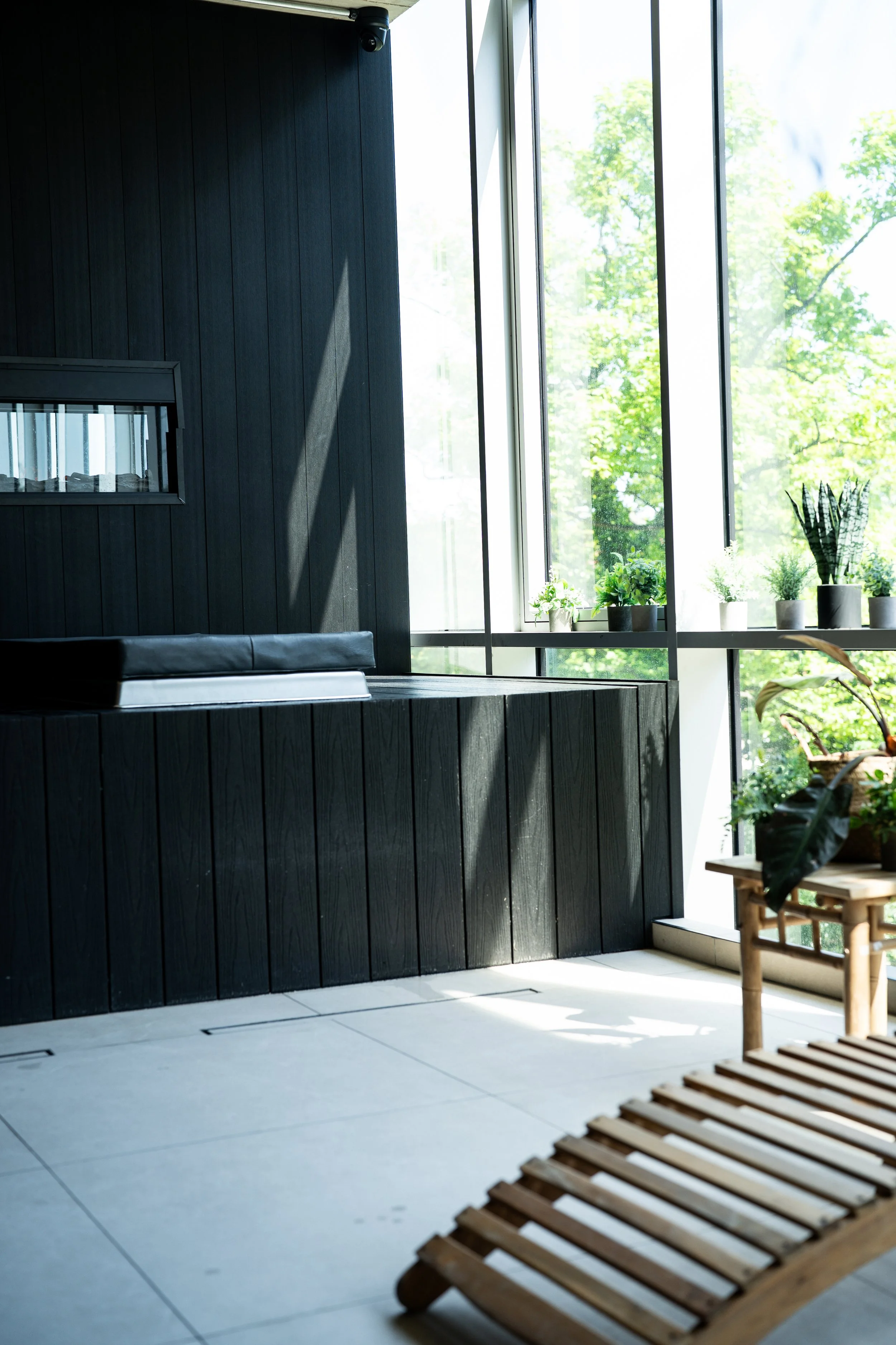 Indoor spa area with large windows, black wooden walls, and potted plants, illuminated by natural sunlight.