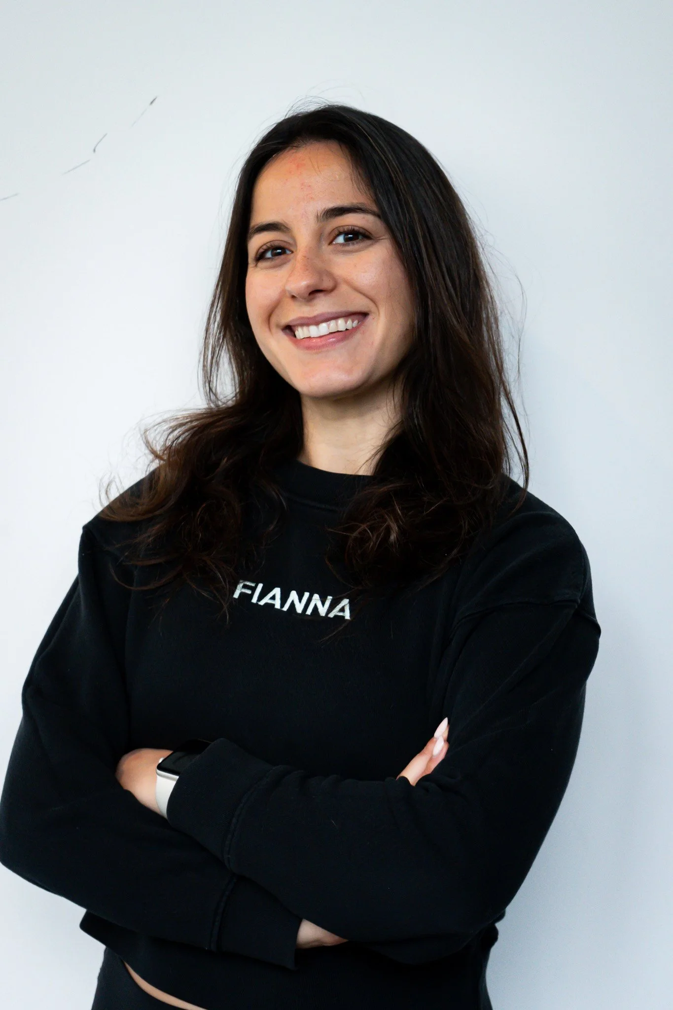Smiling woman with black hair wearing a black sleeveless shirt with the name 'FIANNA' embroidered on it, standing against a black wooden wall.