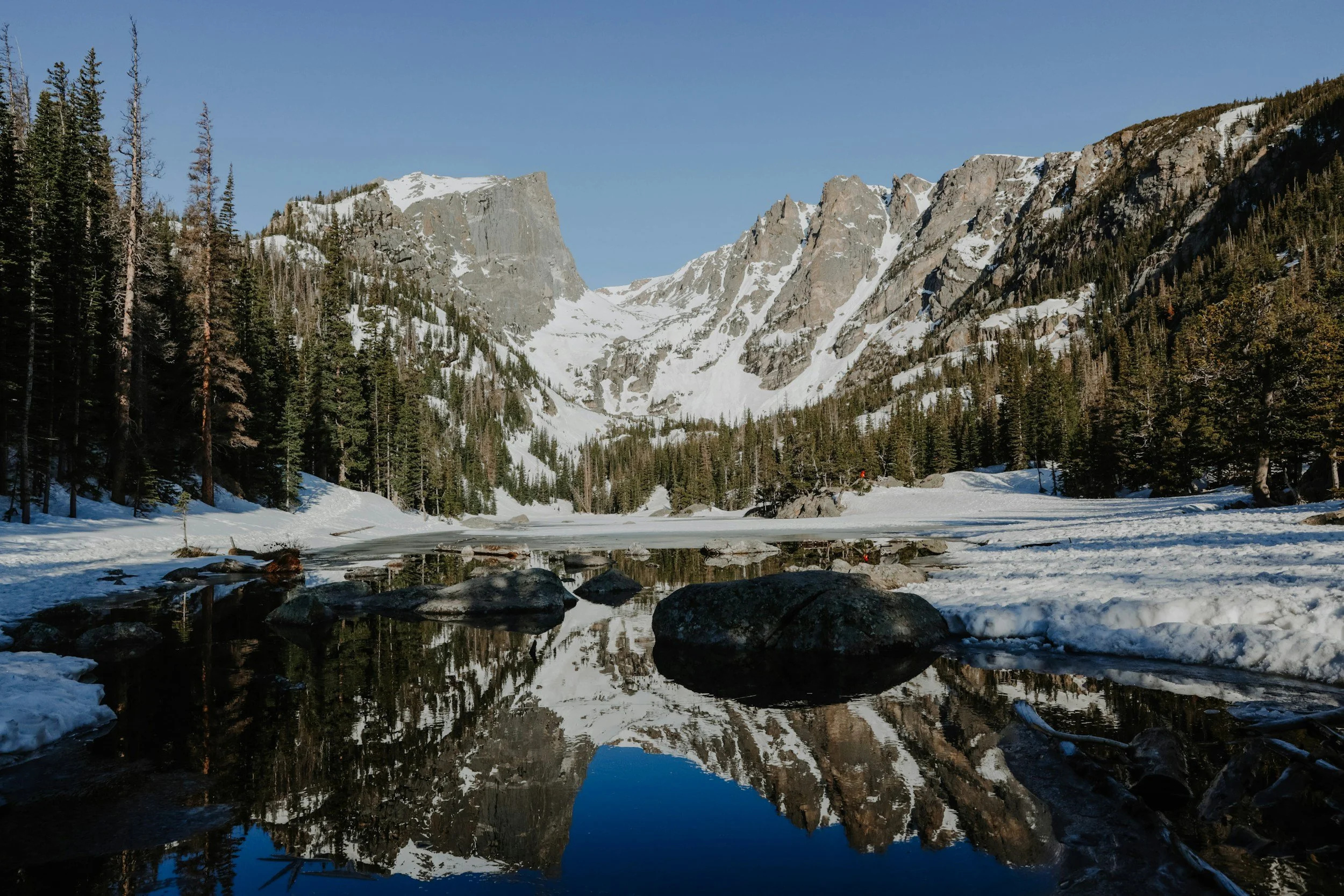 Snow-covered mountains and pine trees reflected in a calm, partially frozen river in a mountainous landscape.