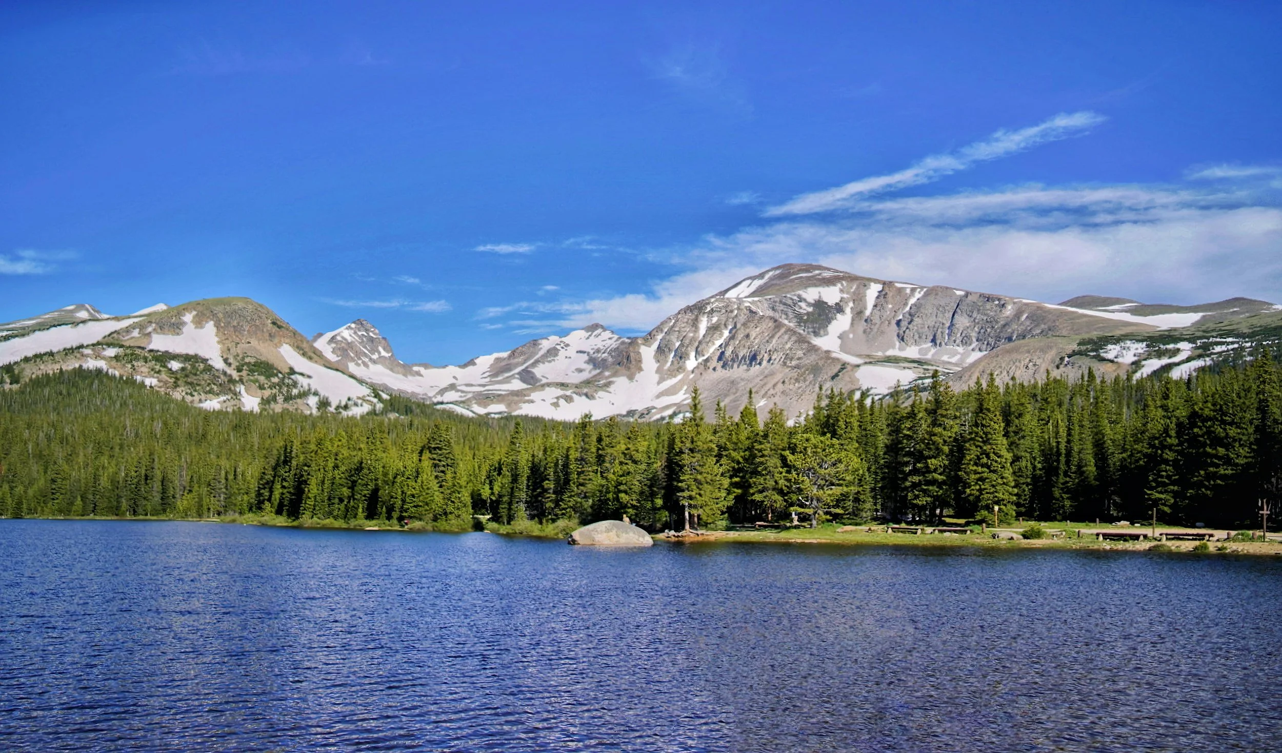 A scenic mountain landscape with snow-capped peaks, green pine forest, and a calm blue lake under a clear blue sky.