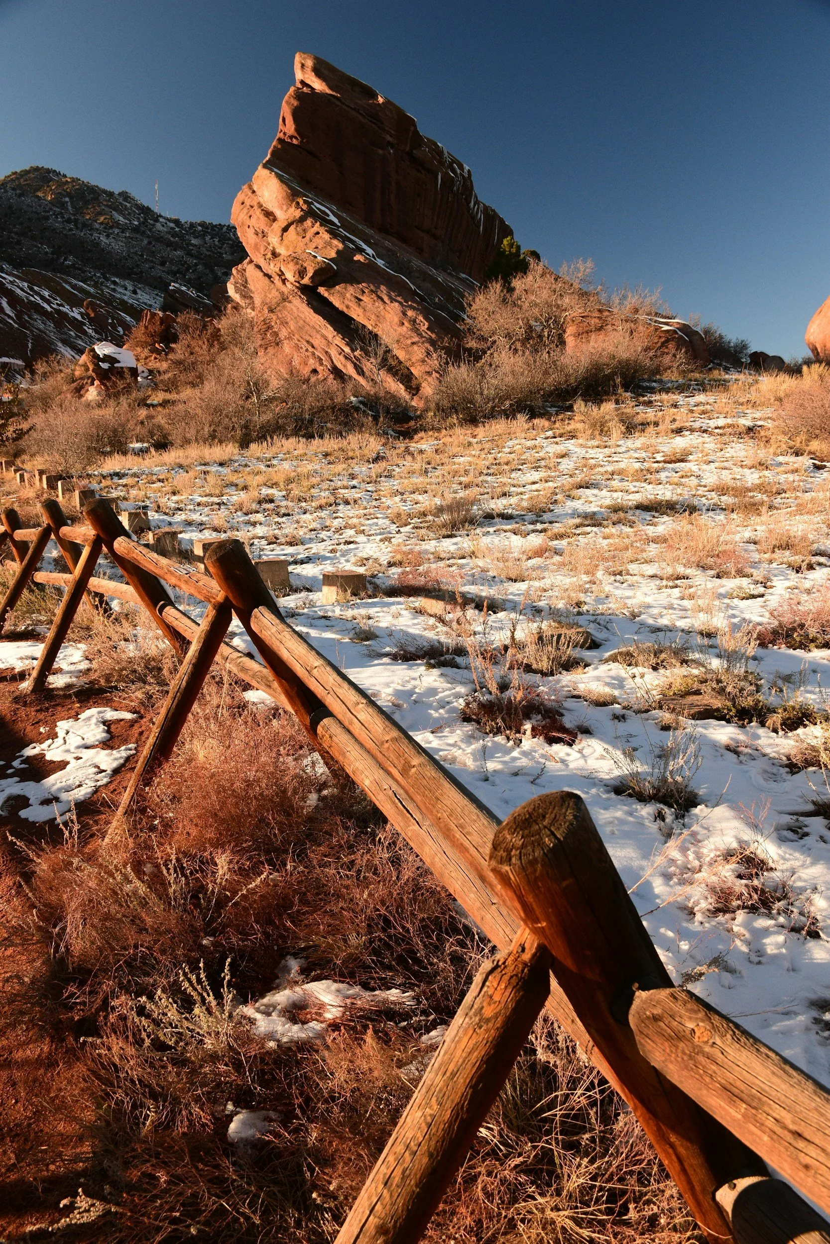 A snow-dusted rocky landscape with a large, tilted red rock formation and a wooden fence along the trail in the foreground.