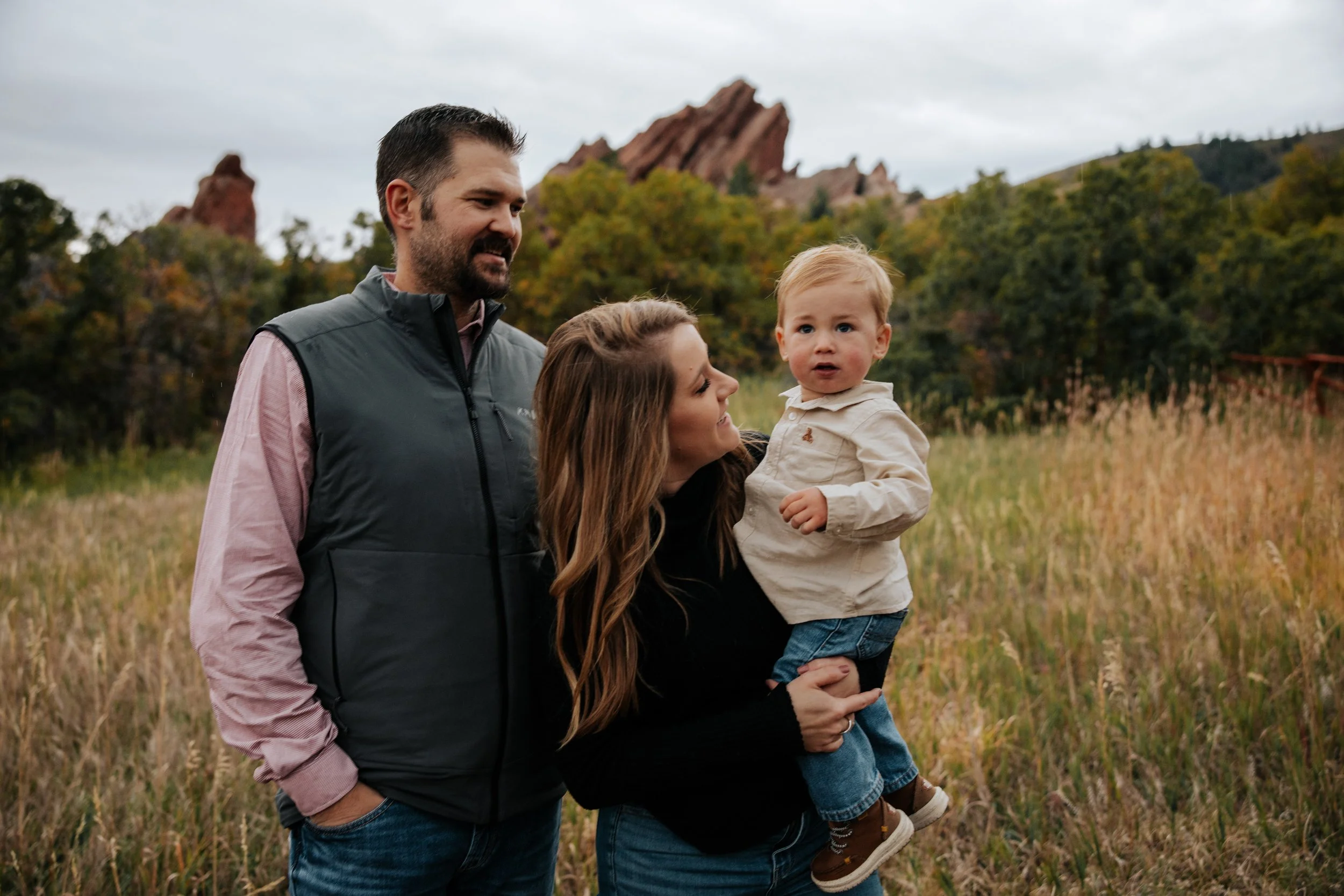 Family of three standing in a grassy field with trees and rocky formations in the background on a cloudy day