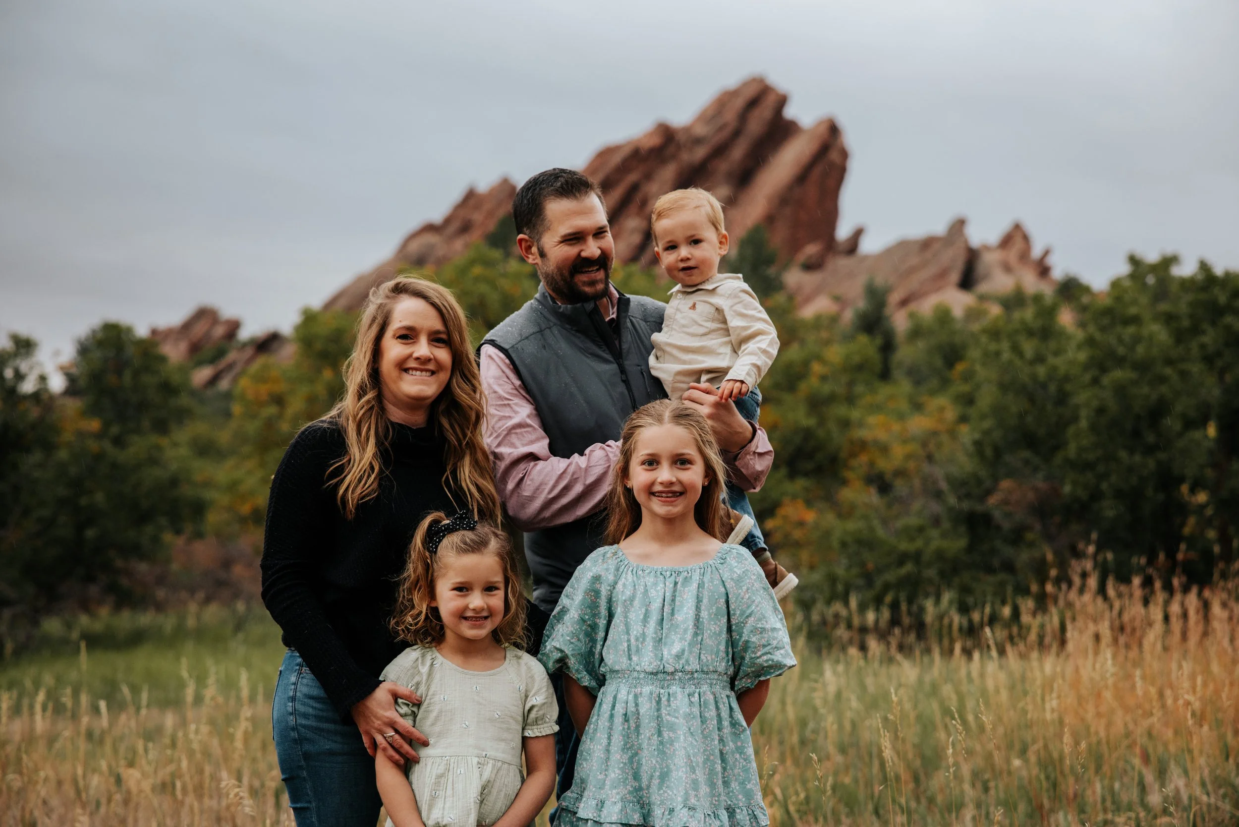 A family of five standing outdoors in a grassy field with trees and rocky formations in the background.