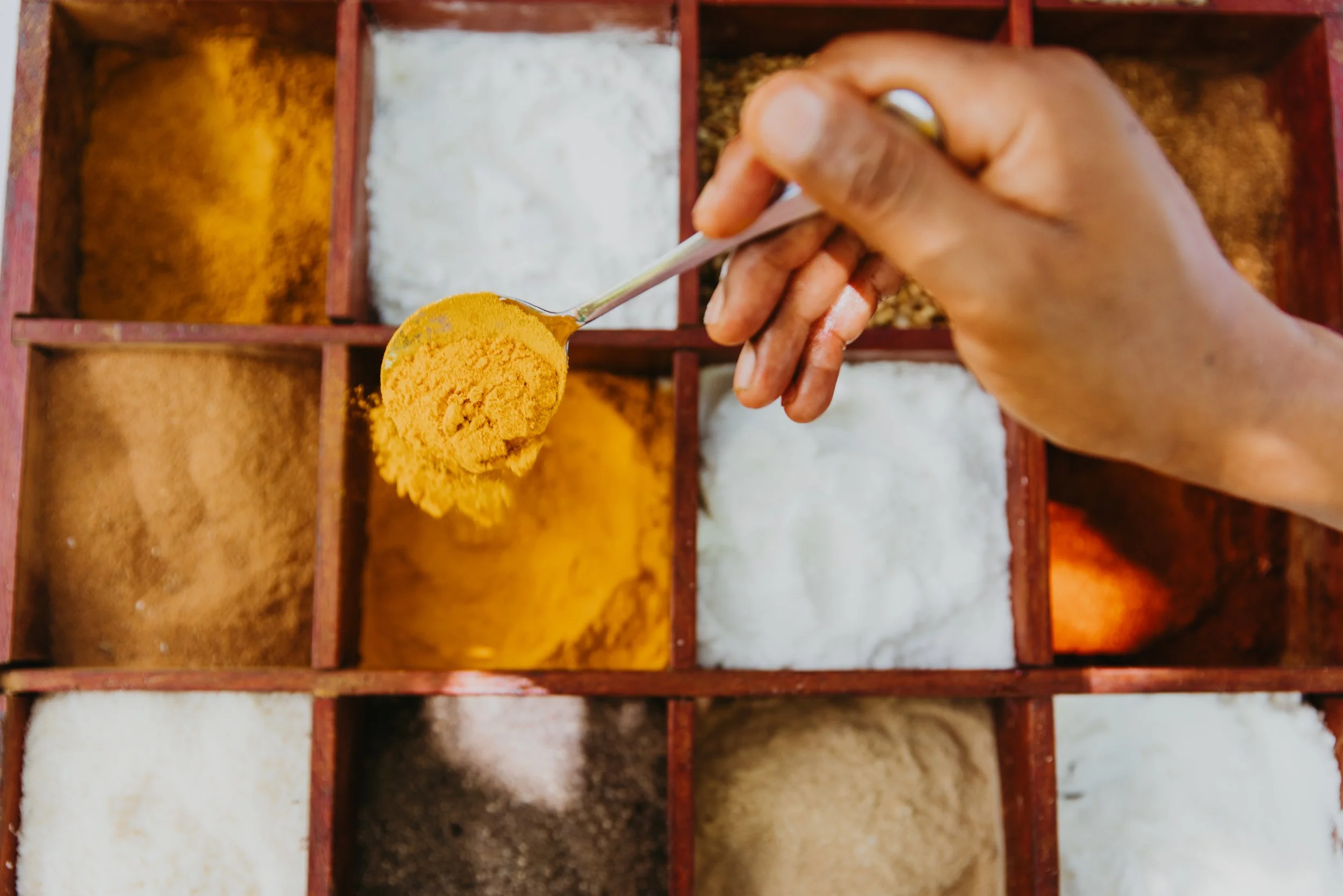 Hand holding a spoon with yellow turmeric powder over a spice box containing various colorful spices in separate compartments.