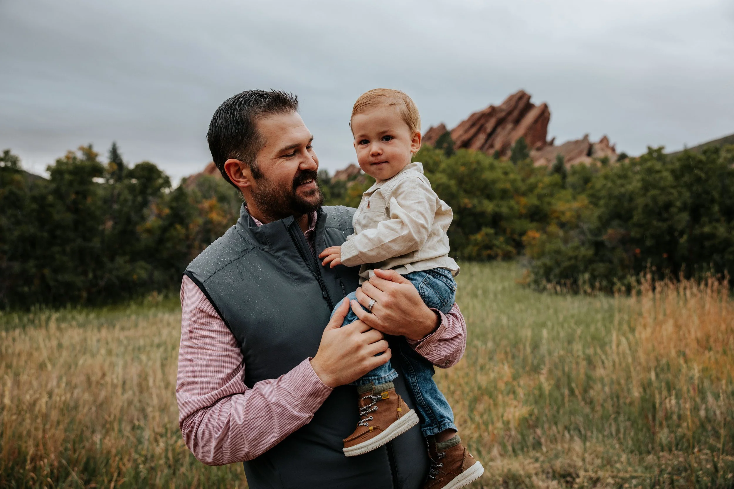 A man holding a young boy outdoors in a grassy field with trees and rocky formations in the background on a cloudy day.