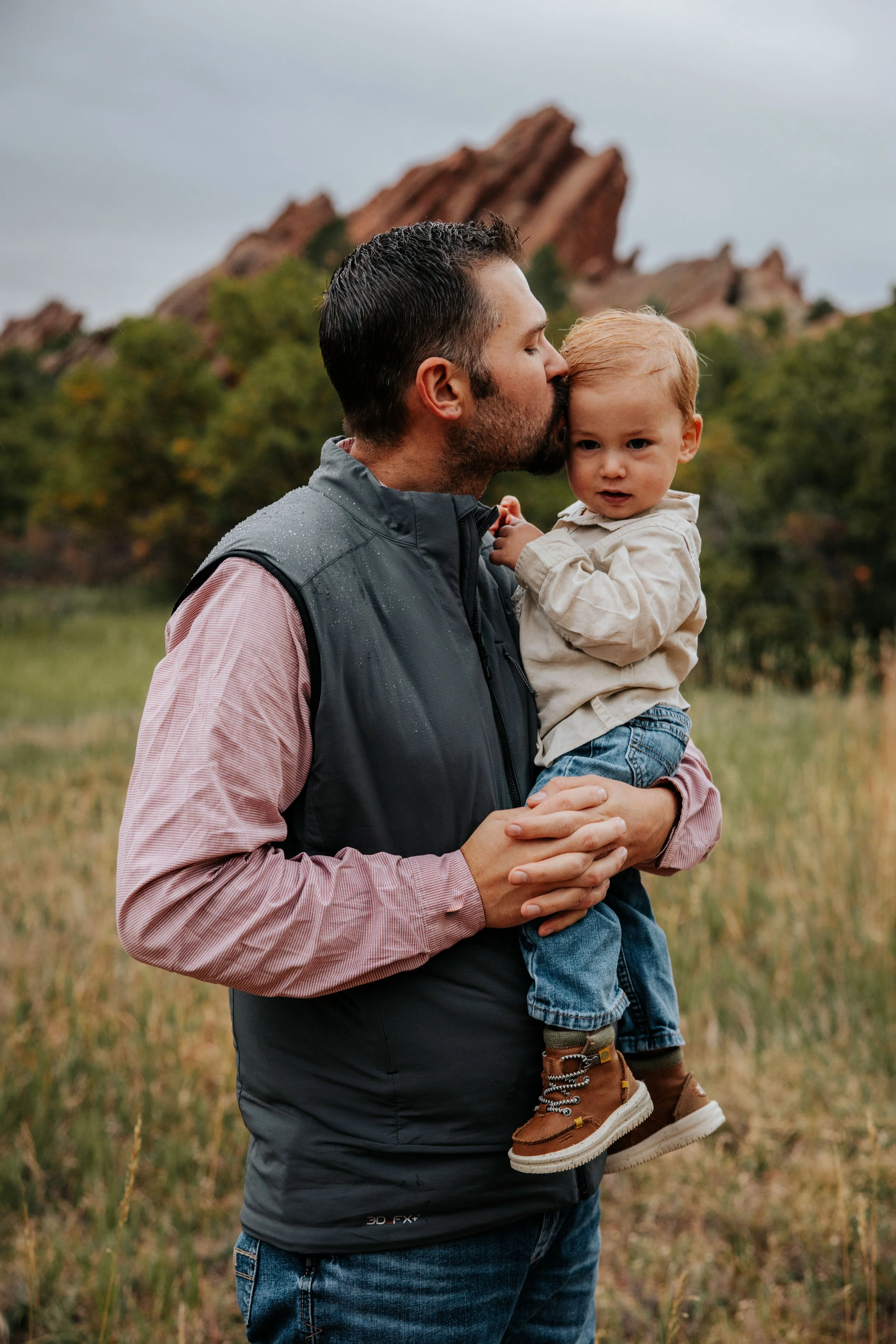 A man holding a young boy in a grassy outdoor area with trees and a rocky mountain in the background.