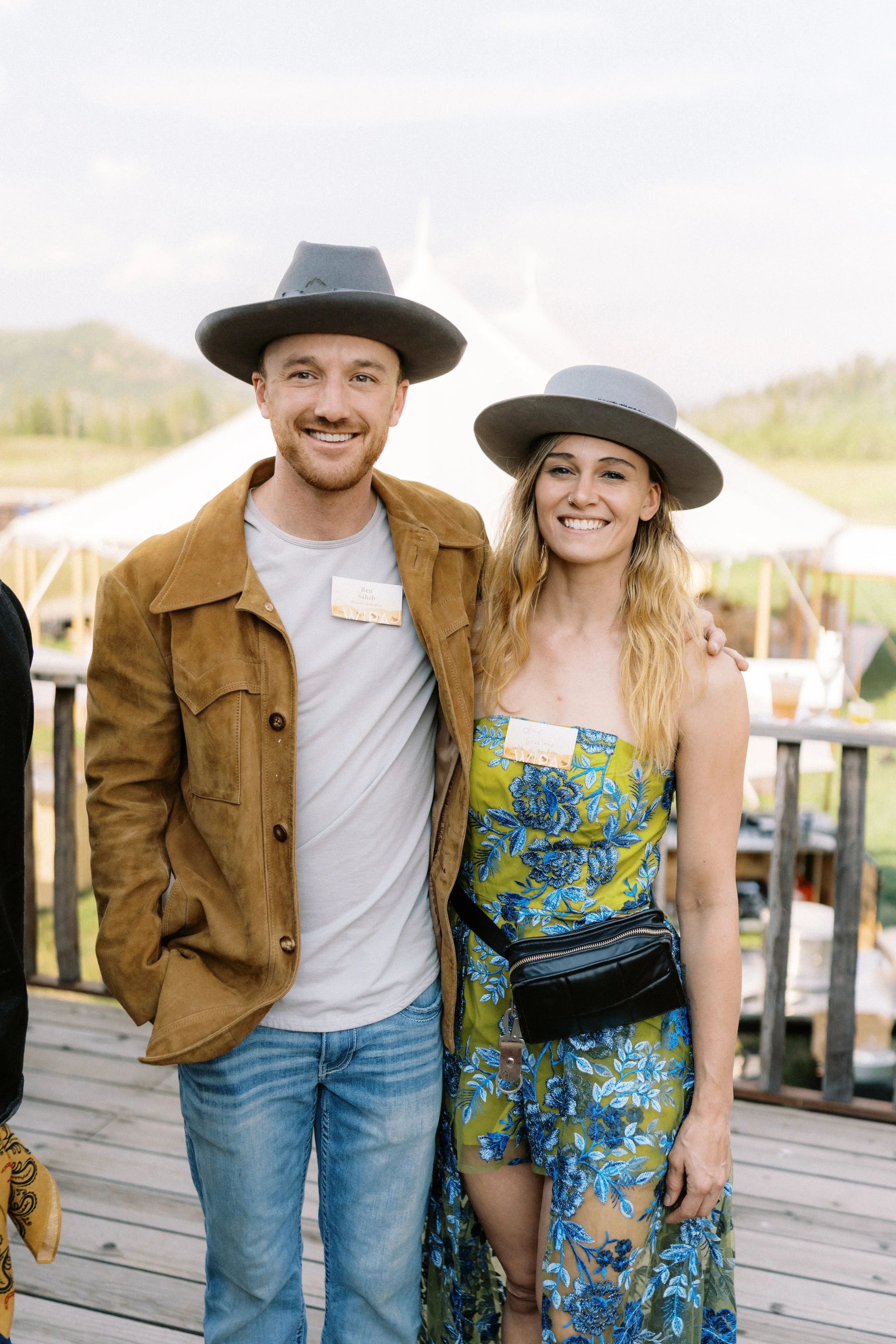 A smiling man and woman posing outdoors with white tents and a wooden deck in the background. The man is wearing a gray hat, light-colored shirt, and brown jacket, while the woman is wearing a gray hat, a green and blue floral dress, and a black waist bag.