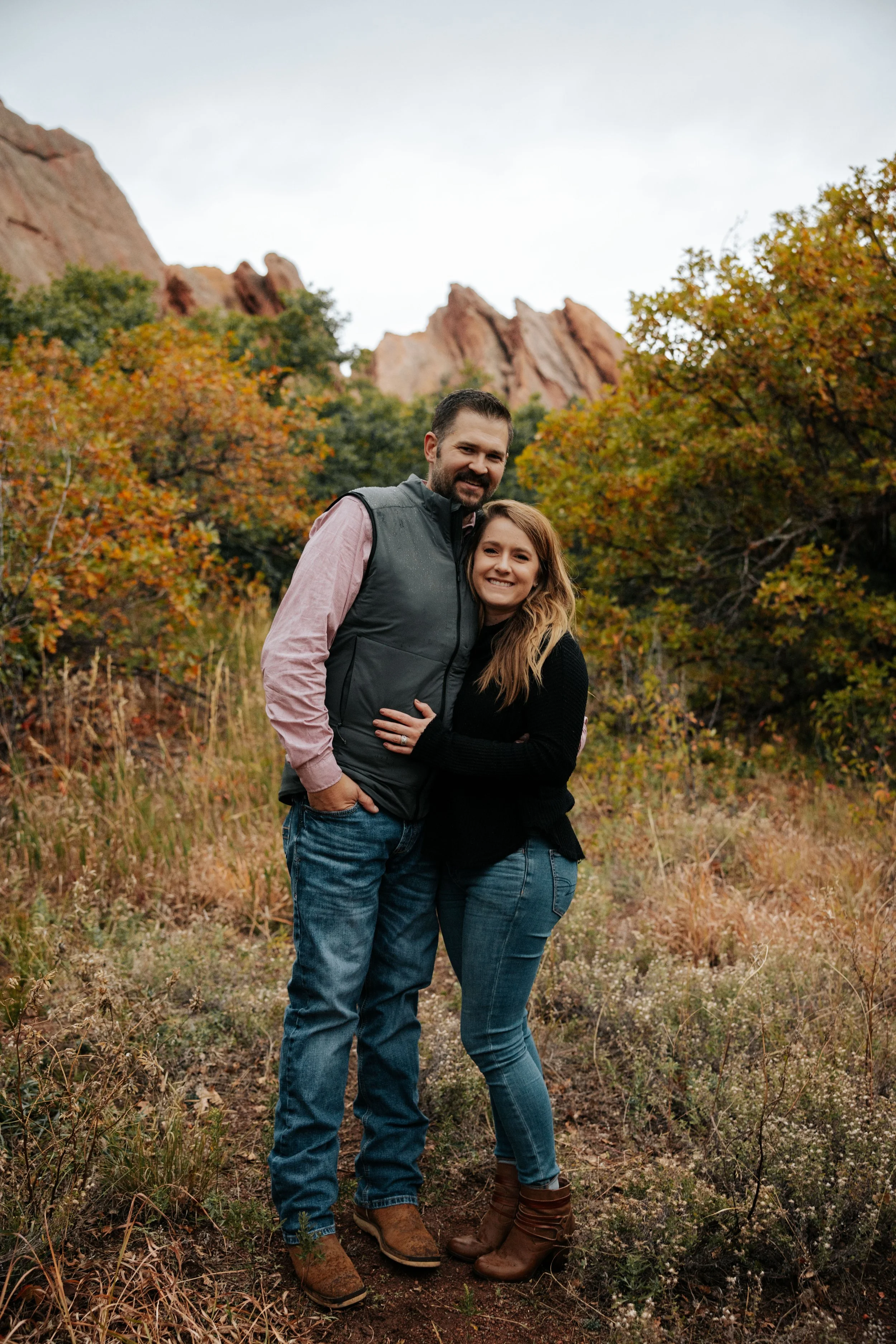 A happy couple standing in a field with autumn foliage and rocky mountains in the background.
