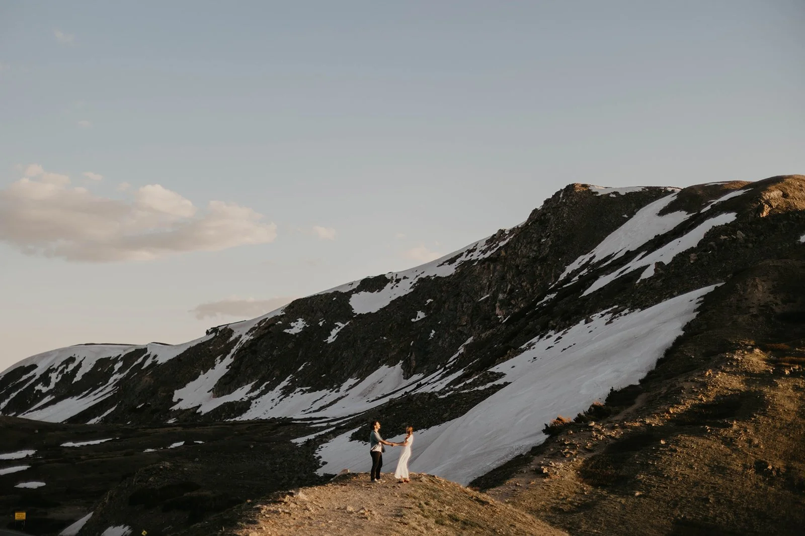 Two people standing on a rocky hill holding hands in front of a mountain landscape with snow patches and a clear sky.