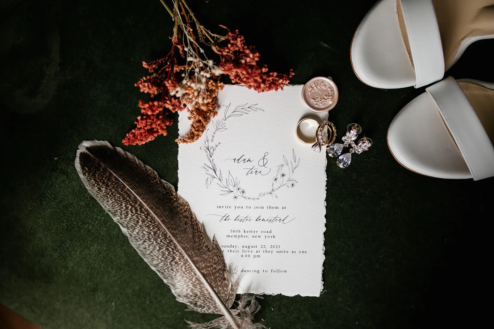 Wedding invitation surrounded by a feather, dried flowers, a ring, earrings, and an ink stamp, with a pair of white shoes nearby.