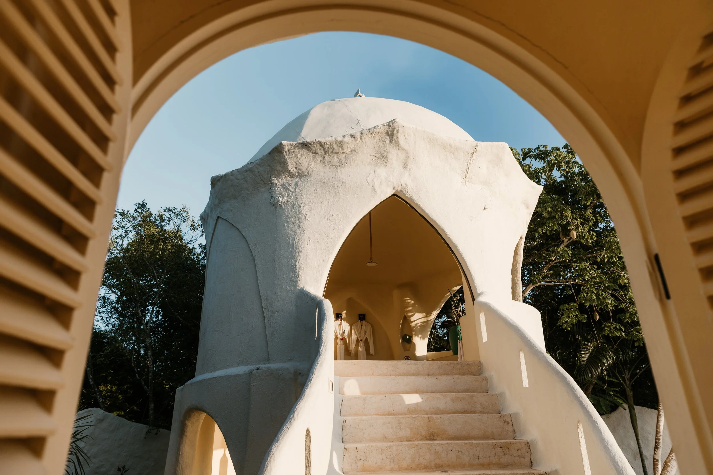 Dome-shaped architectural structure with an archway entrance and staircase, surrounded by trees, viewed through a wooden louvered frame.