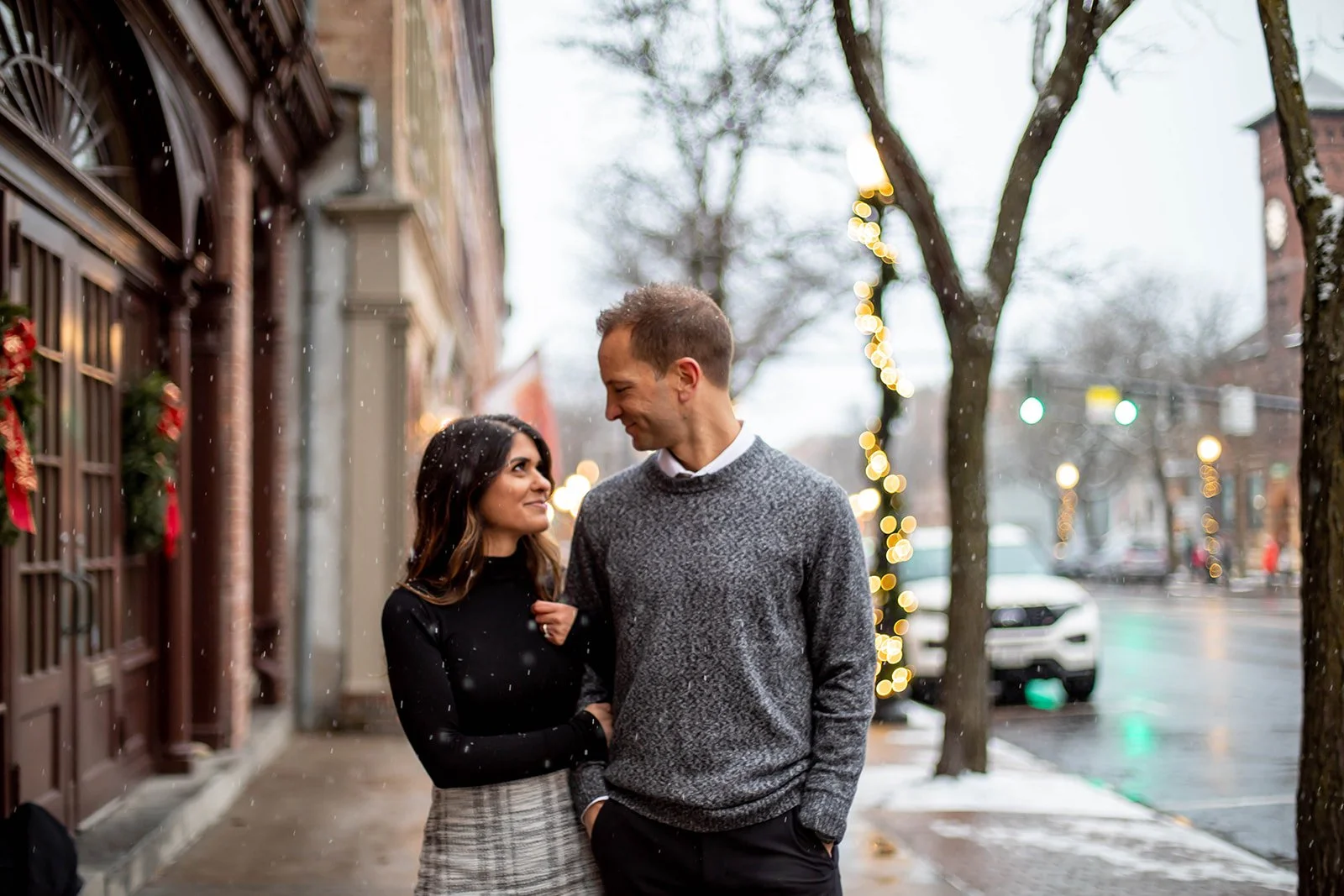 A couple standing on a city sidewalk during snowfall, looking at each other. The woman has long dark hair and is wearing a black top and a plaid skirt. The man has short hair and is wearing a gray sweater. Christmas decorations and string lights are 