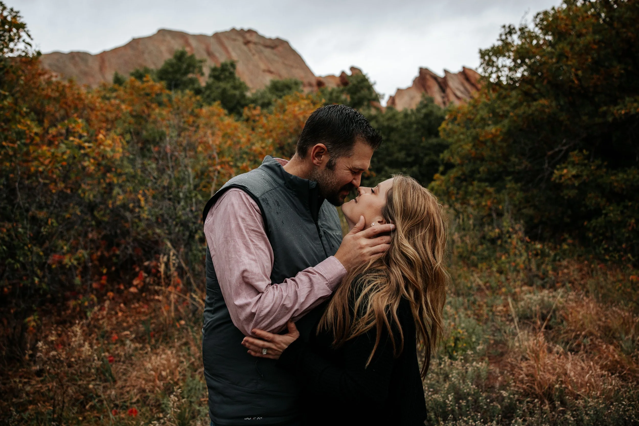 A man and woman embrace in an outdoor setting with autumn foliage and mountains in the background.