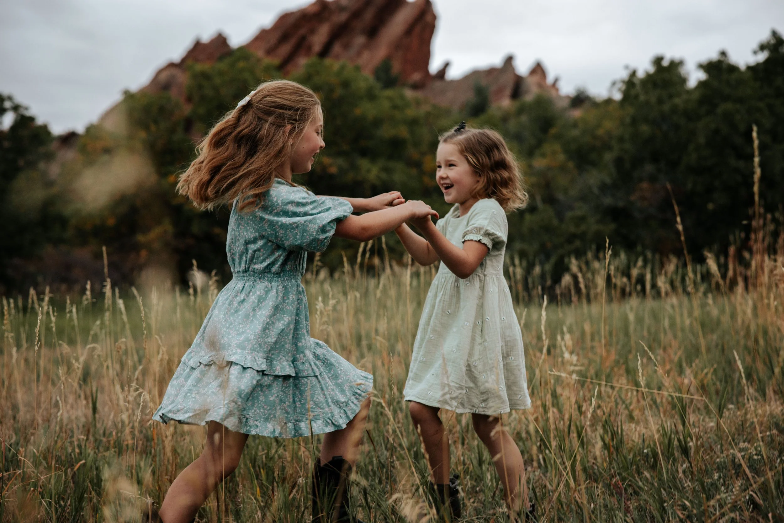 Two young girls are playing and laughing together in a grassy field, holding hands and spinning around, with trees and rocky hills in the background.