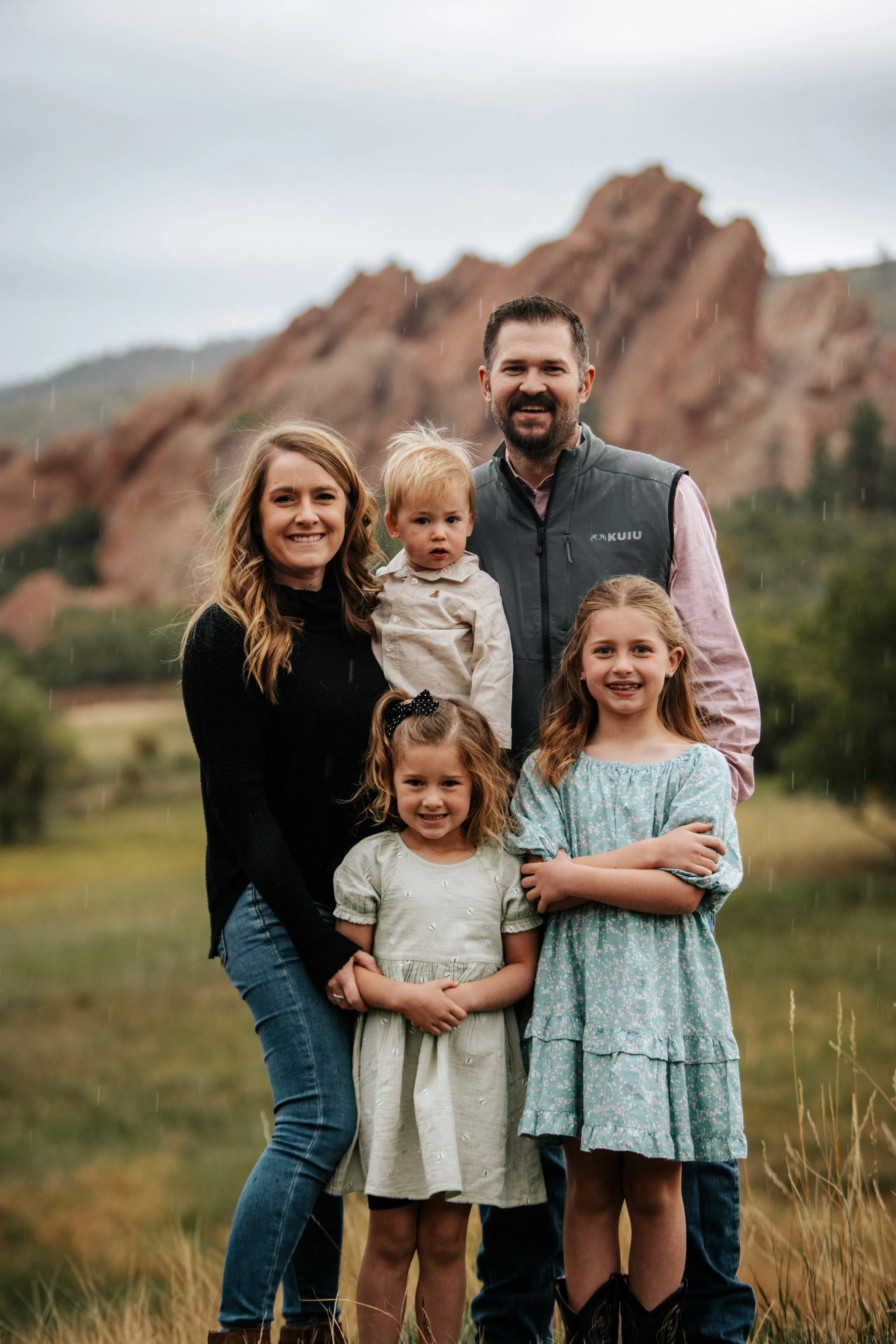 Family of six, including two Adults, one teenage girl, two young girls, and a young boy, standing outdoors in a grassy area with a mountain and cloudy sky in the background, rainy weather.