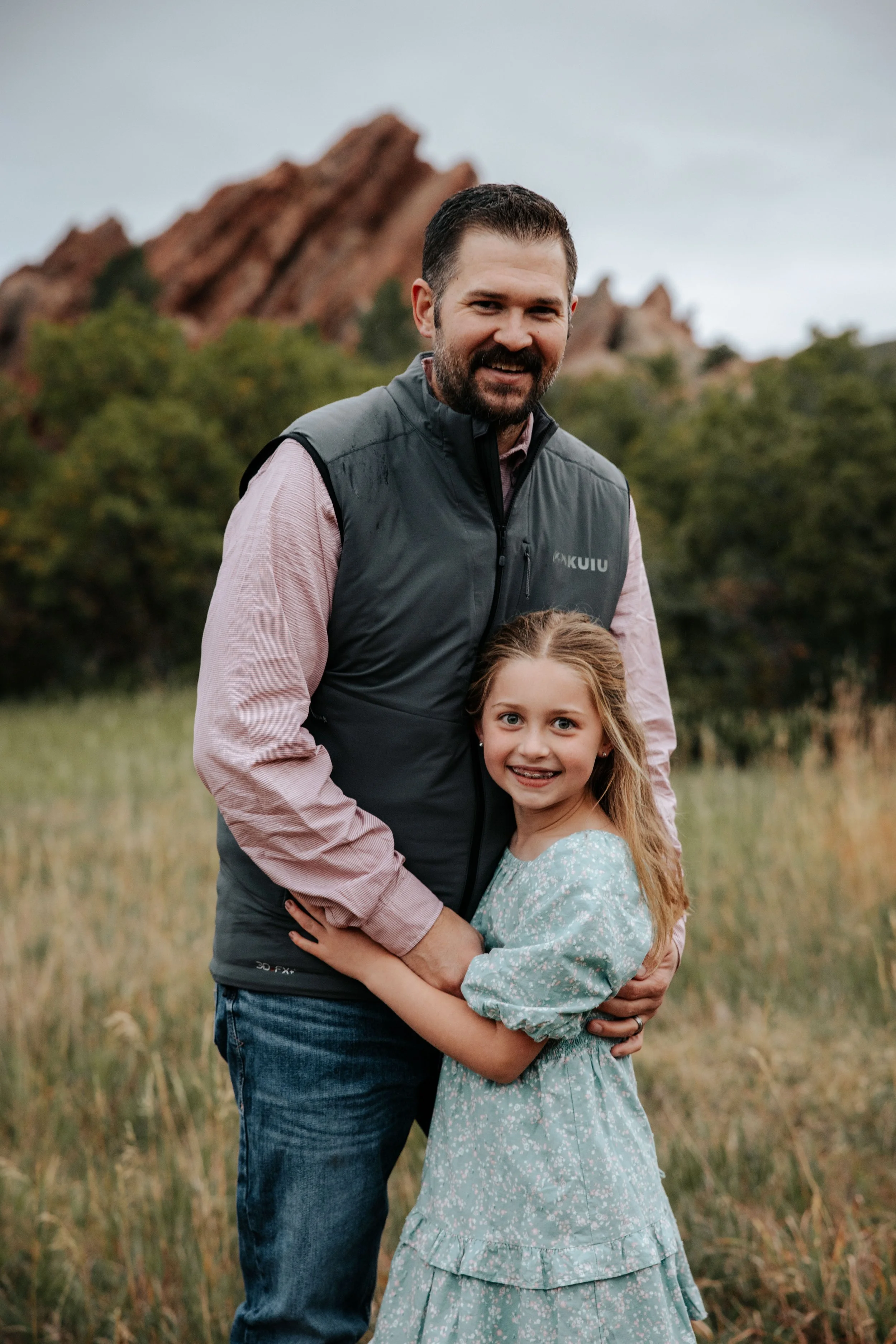 A man and a young girl standing outdoors in a grassy area with trees and red rock formations in the background, smiling and embracing.