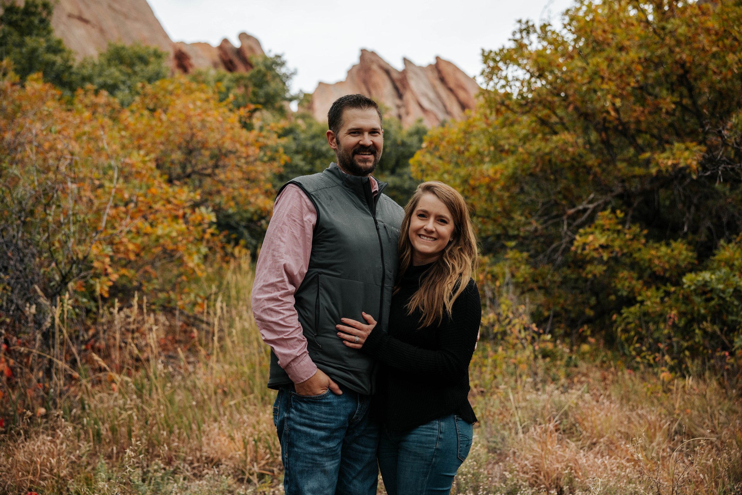 A smiling couple standing outdoors in a fall landscape, with orange and yellow trees and rocky formations in the background.