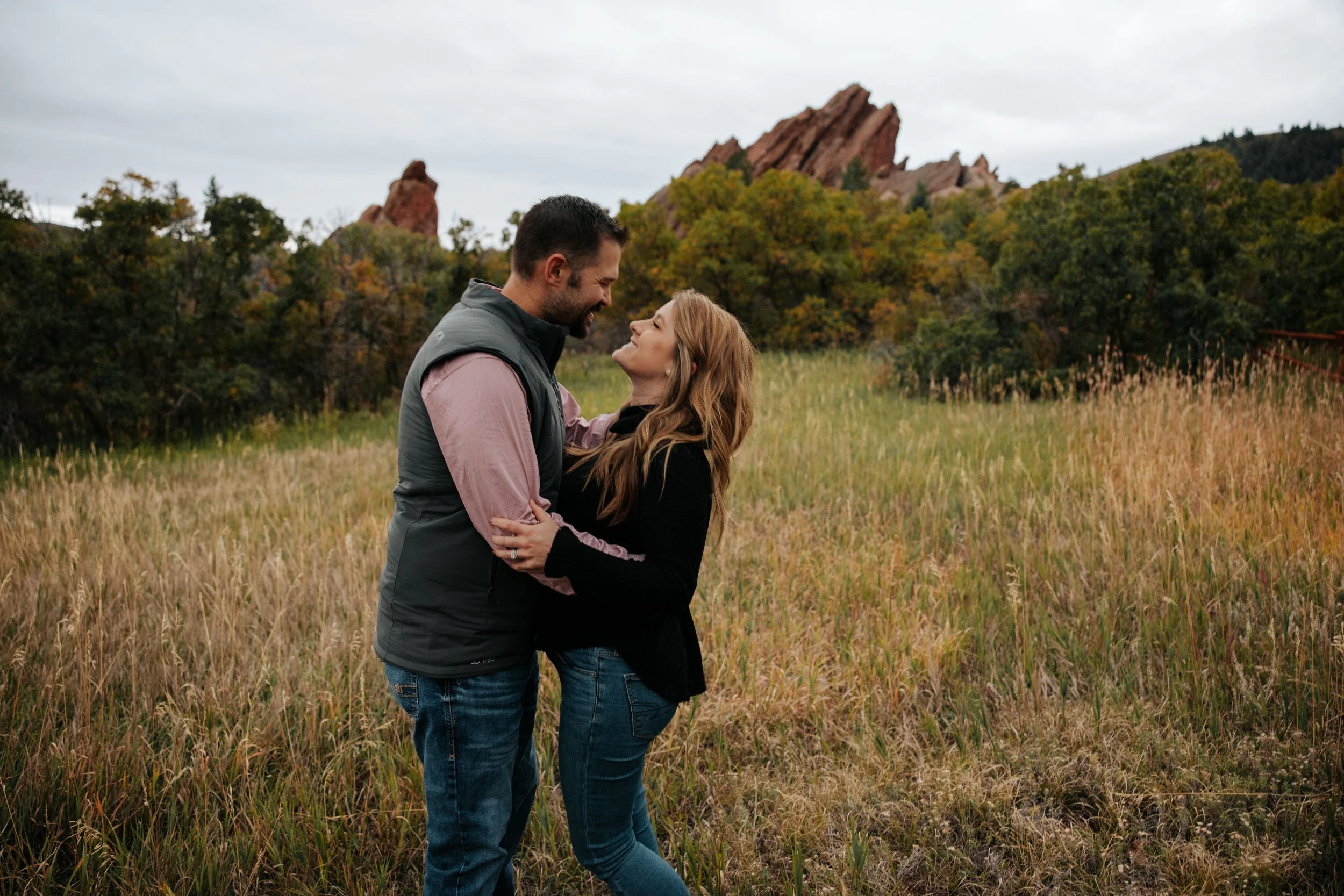 A couple standing close together in a grassy field, looking at each other affectionately, with a backdrop of trees and red rock formations under an overcast sky.