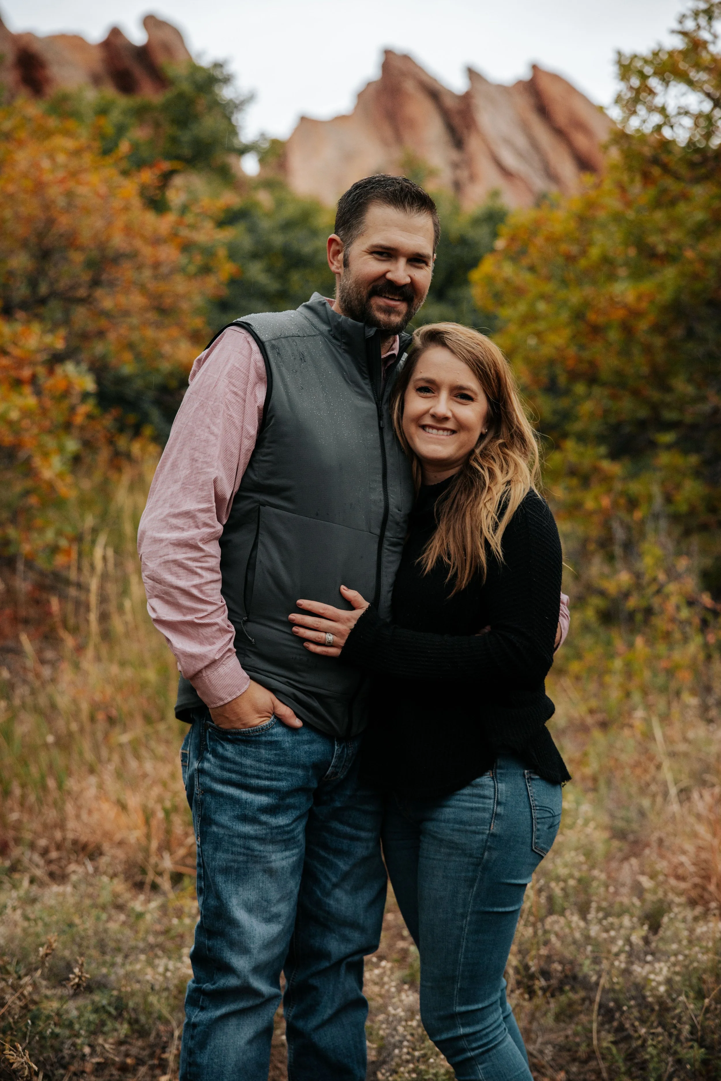 A smiling couple standing outdoors in a forested area with colorful autumn foliage and rocky formations in the background.