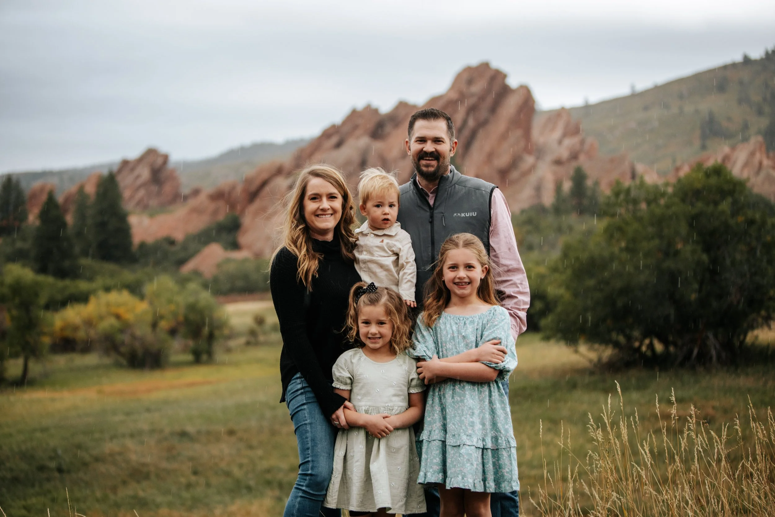 Family of six standing outdoors in a grassy field with mountains and trees in the background during rainy weather.