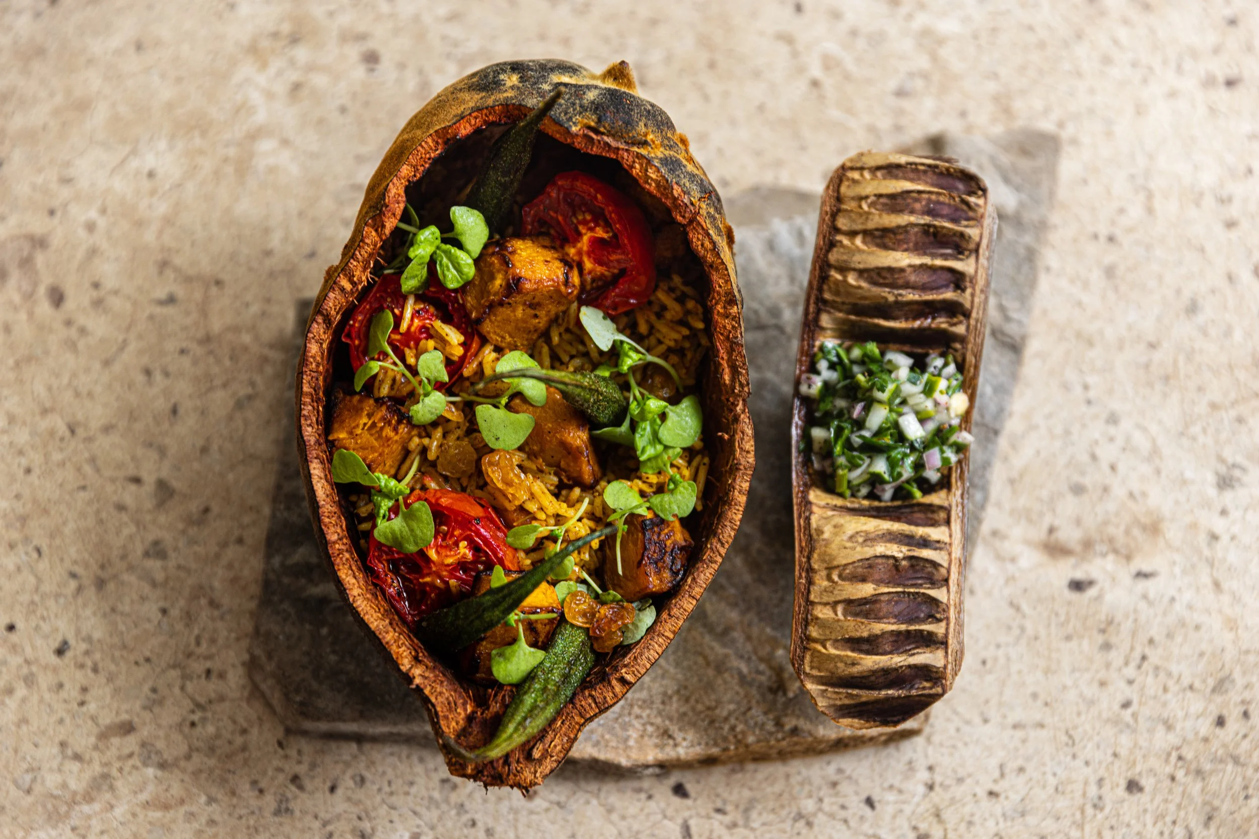 A gourmet dish served in a dried gourd bowl, featuring rice, grilled vegetables, microgreens, and herbs. Beside it, there is a small wooden platter containing a chopped herb garnish.